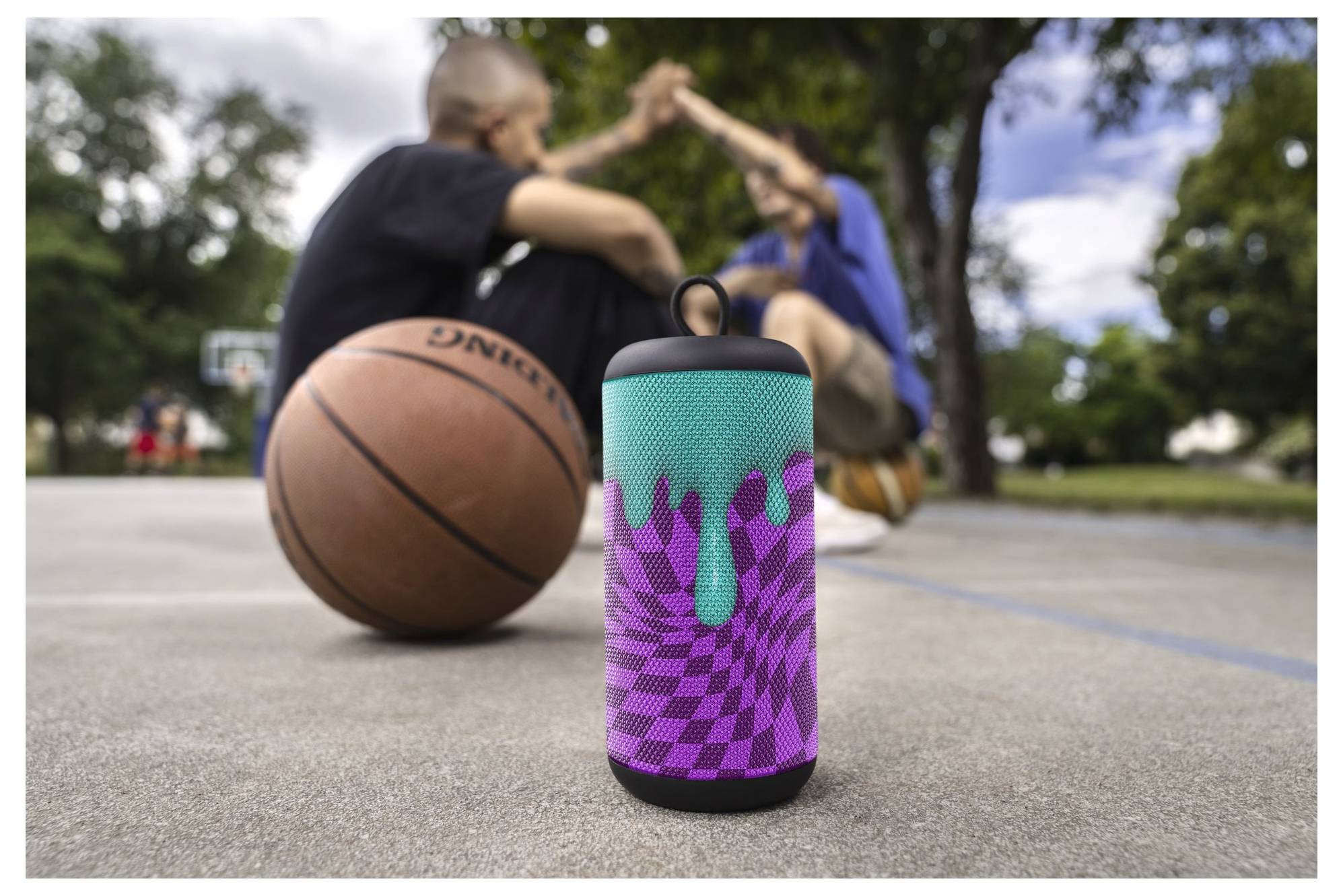 A colorful portable speaker is placed on a basketball court. Two people are sitting and interacting in the background near a basketball.