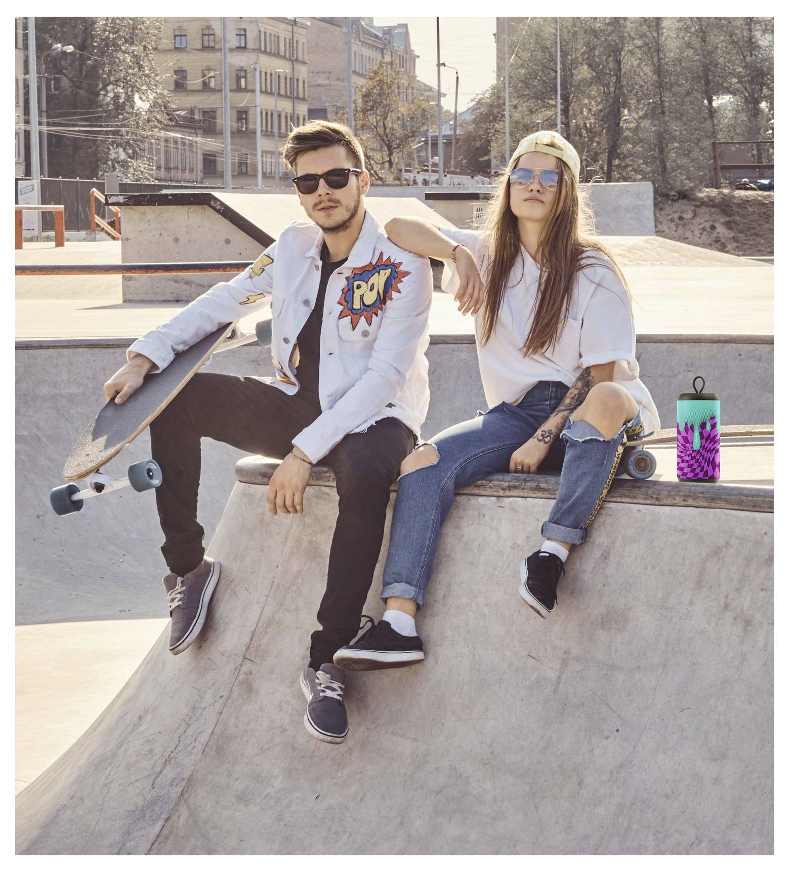 Two individuals sitting on a concrete skate park structure. One holds a skateboard. Both wearing casual clothes and sunglasses.