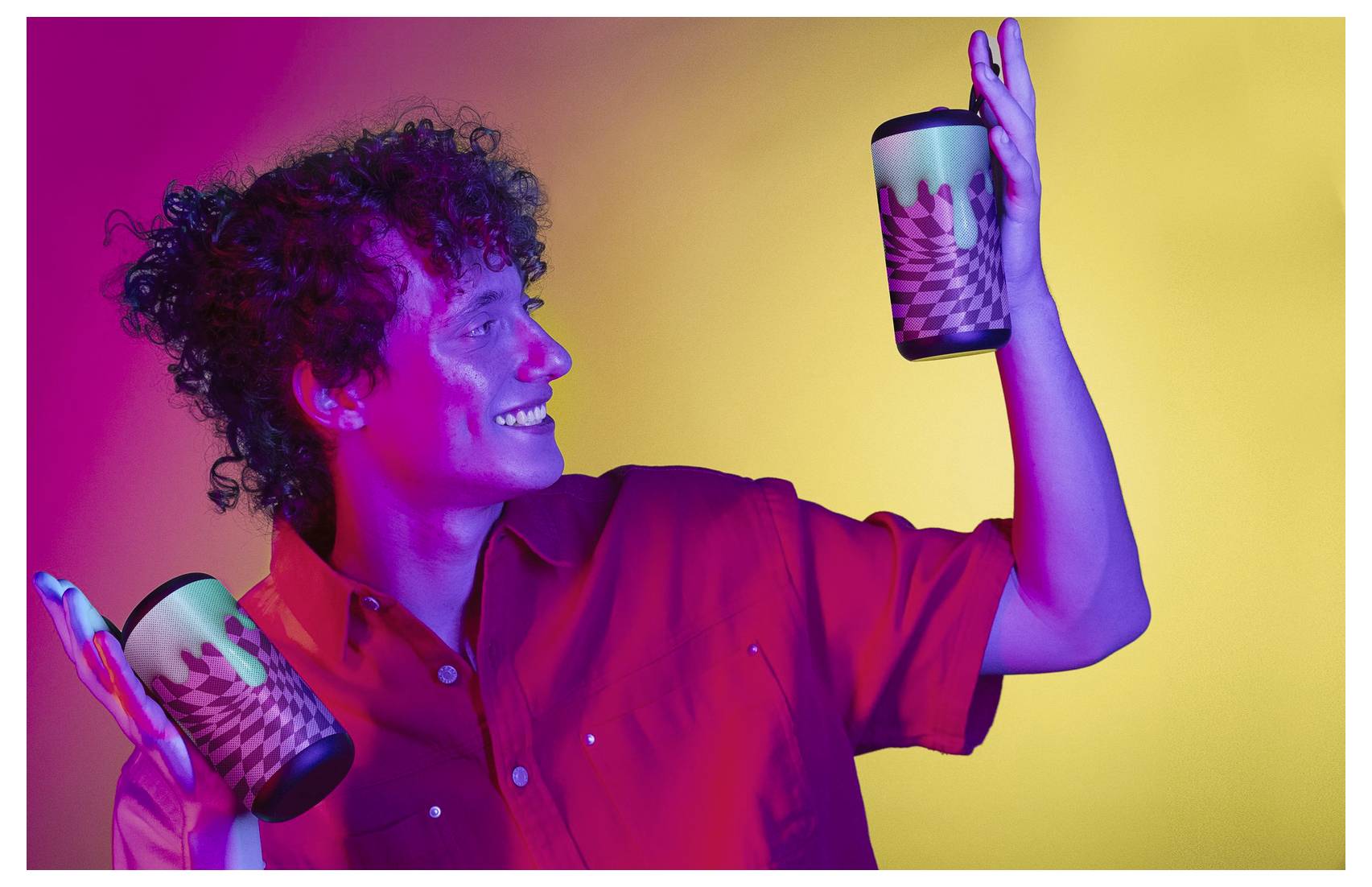 A person in a red shirt smiles while holding two colorful cups, set against a vibrant purple, red, and yellow background.