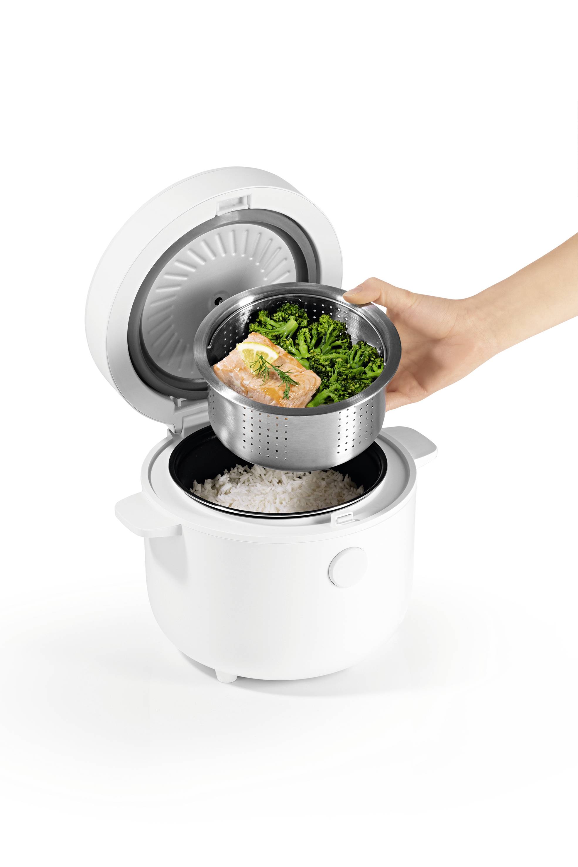 A person is placing a steaming tray with cooked salmon and greens into a rice cooker, which already has rice inside.