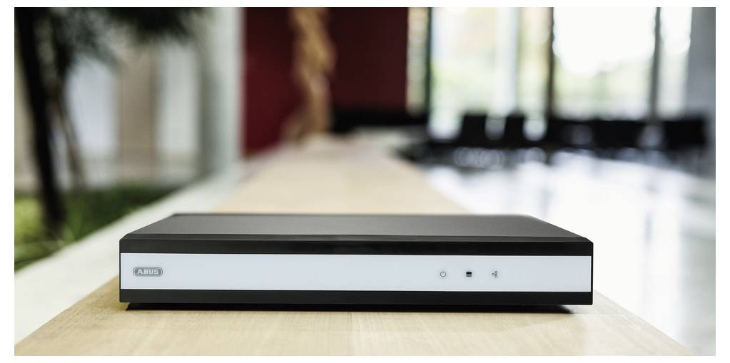 A black electronic device, possibly a media player or modem, sits on a wooden surface in a modern, well-lit room with large windows.