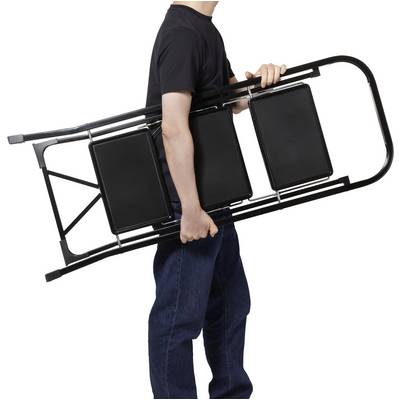 Person carrying a folded black folding chair, wearing a black shirt and blue jeans, against a plain white background.