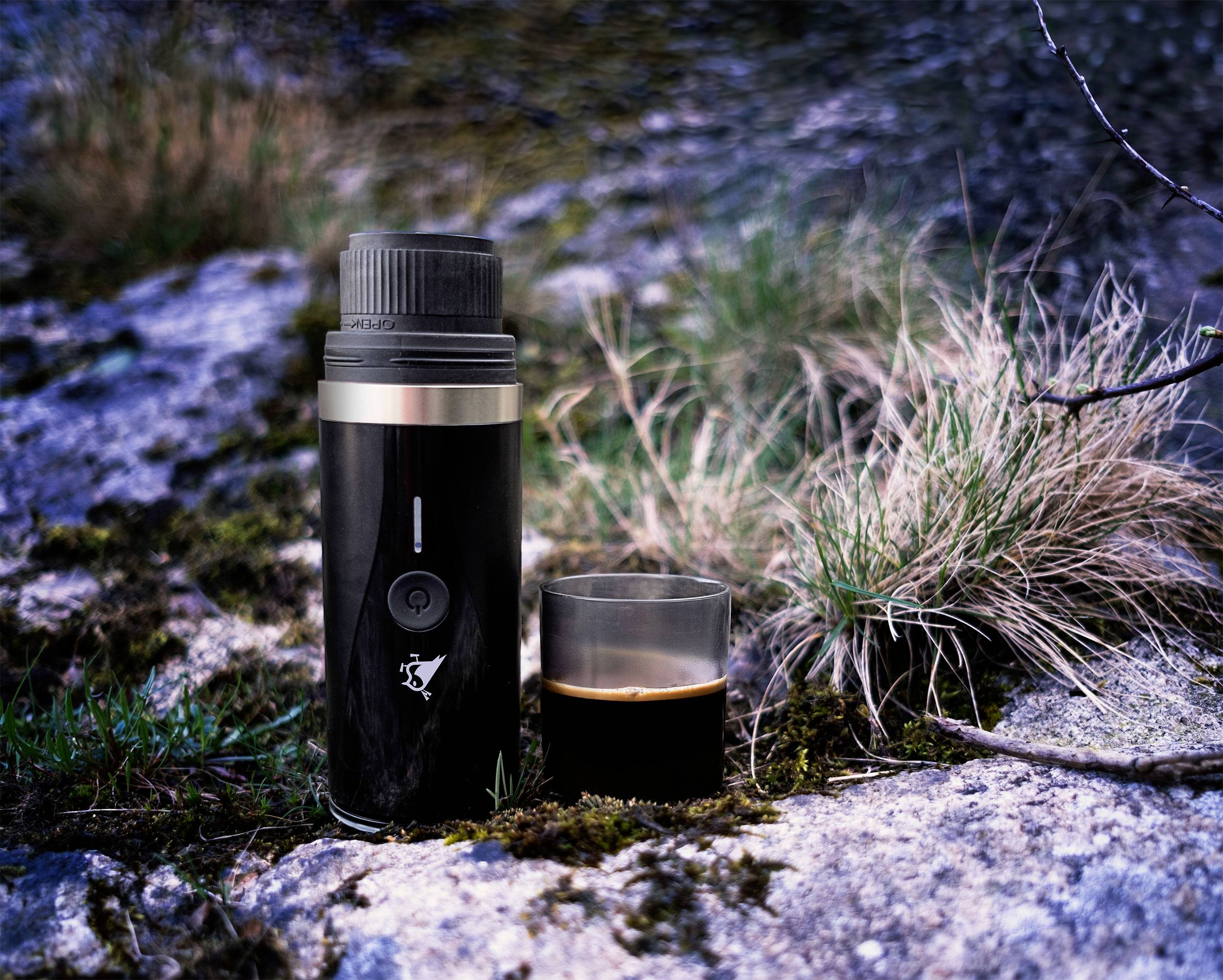 A black thermos and a half-filled cup of coffee sit on a mossy rock, surrounded by grass and rocky terrain.