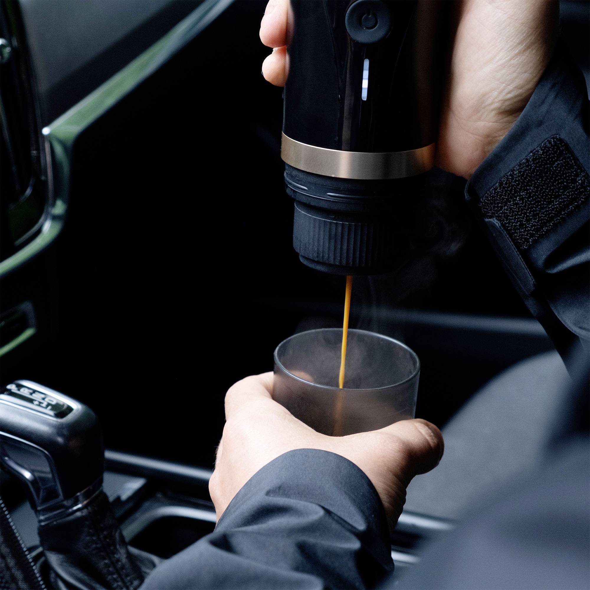A person is pouring coffee from a travel mug into a small cup inside a car. The mug is black with a gold band near the top.