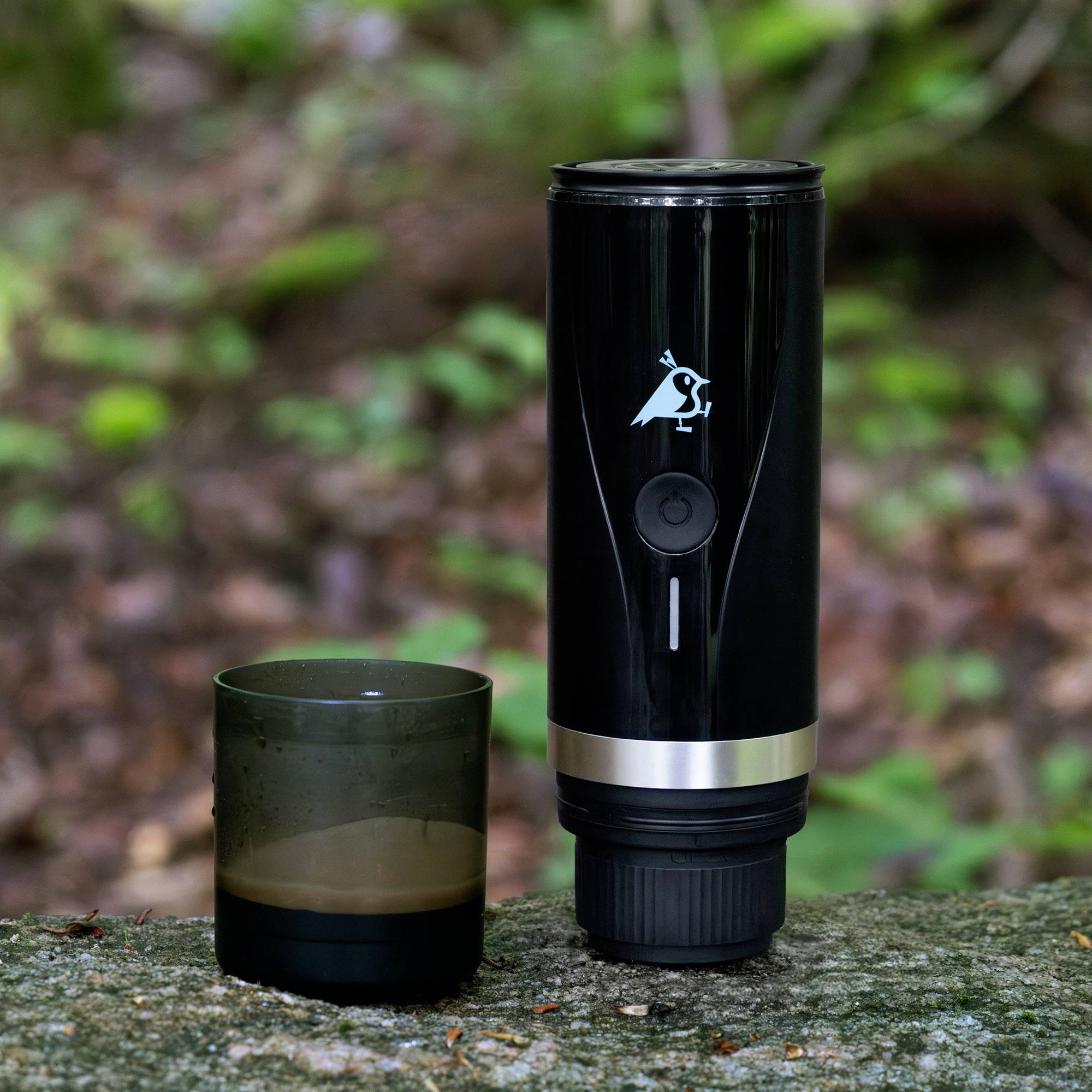 'Black portable coffee grinder with an owl logo, next to a dark cup filled with coffee, set on a stone surface with a blurred forest background.'