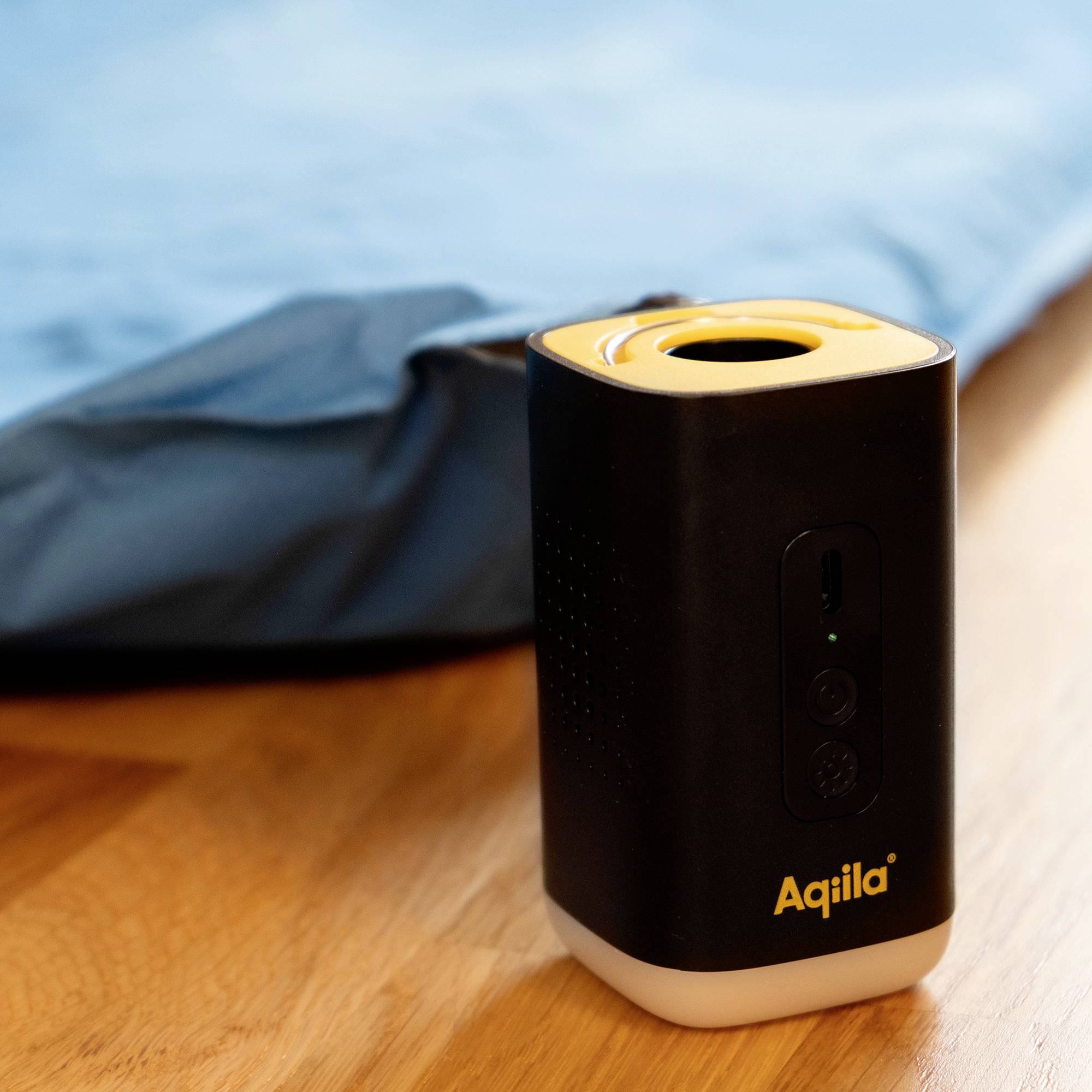 Black rectangular electronic device with yellow top, showing logo 'Aqila' at the base. Positioned on a wooden surface near blue fabric.