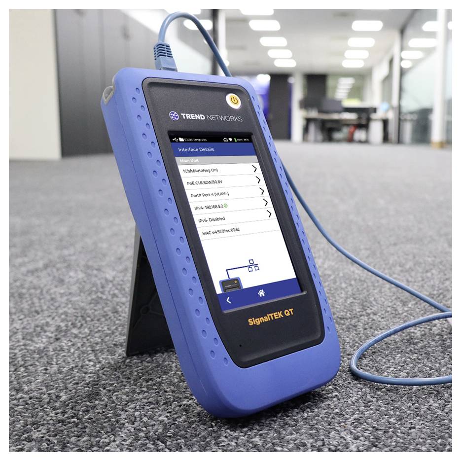 A network testing device on a carpeted office floor shows interface details on its screen with a connected blue cable.