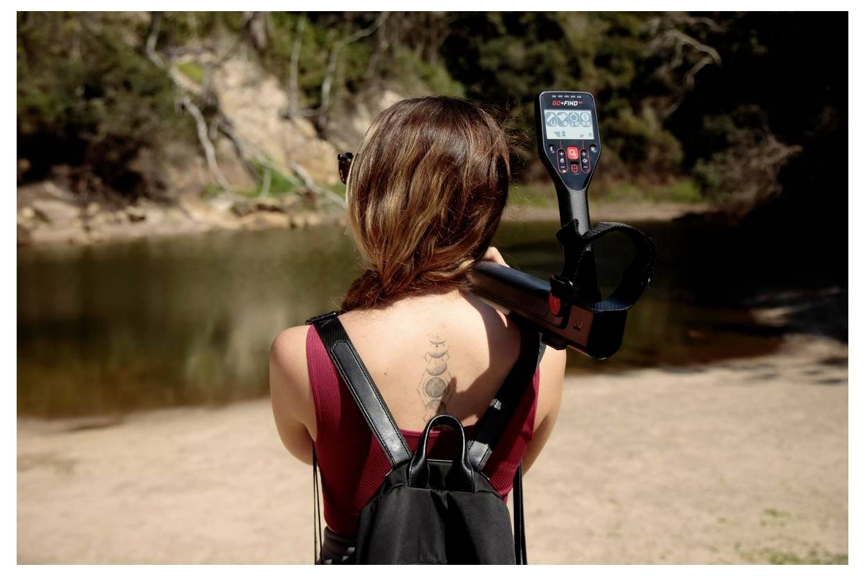 A person with long hair stands near a tranquil lake, holding a metal detector over their shoulder, facing the water surrounded by trees.