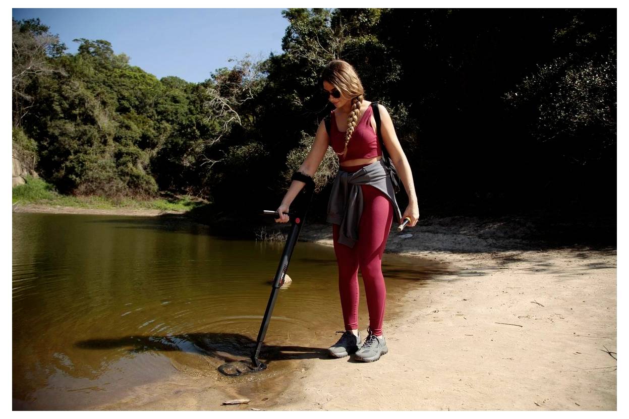 A person wearing activewear stands by a lakeshore, using a walking stick to inspect the water. Trees and foliage are visible in the background.