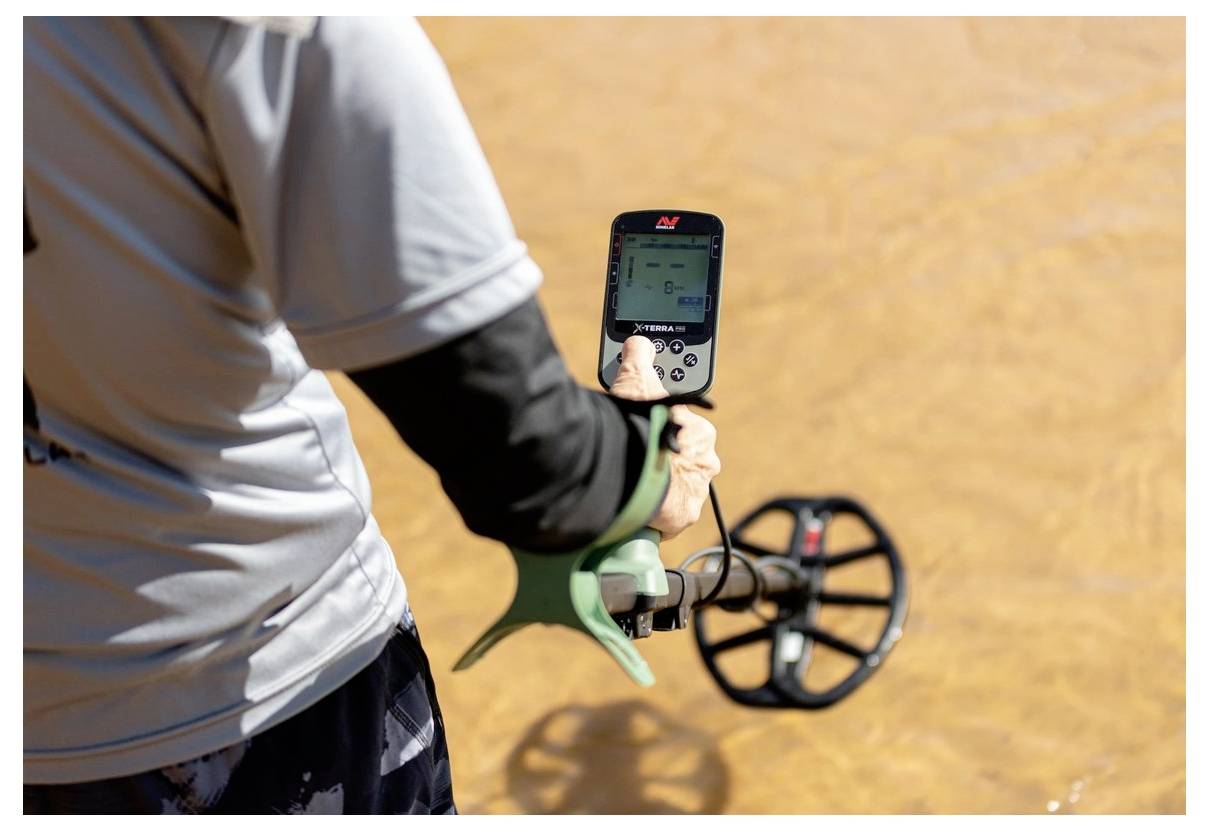 A person uses a metal detector on brown, muddy ground, focusing on the device's screen.