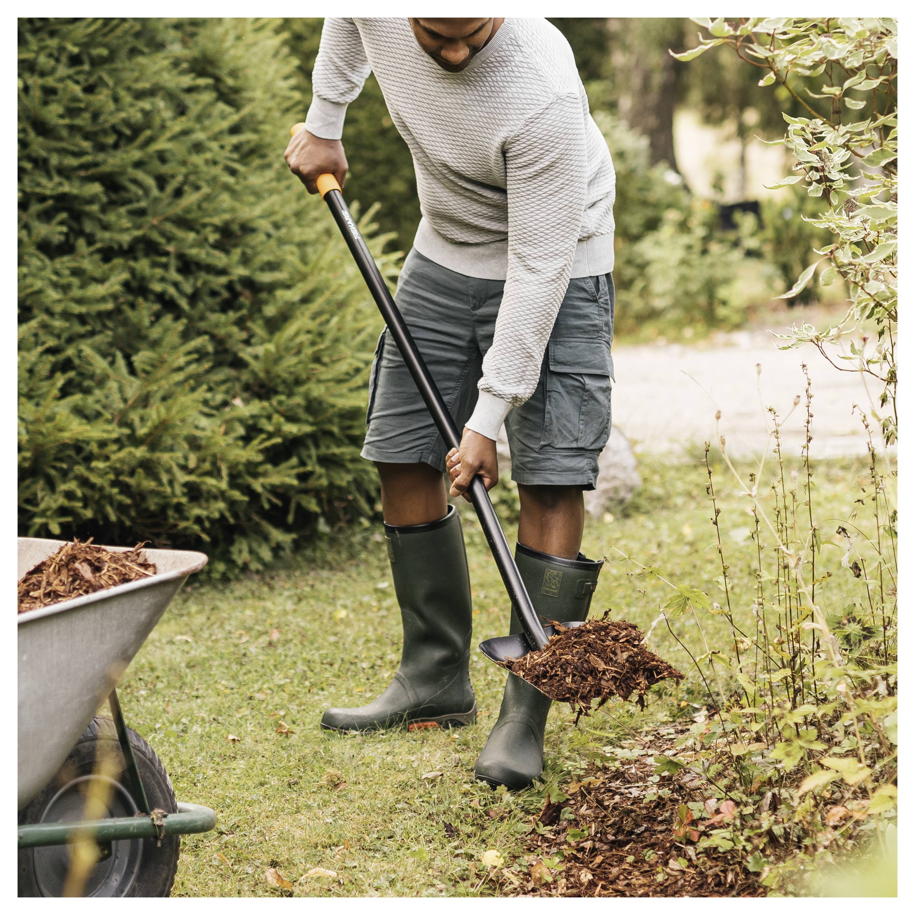 A person wearing a sweater, shorts, and boots uses a shovel to transfer mulch from a wheelbarrow to a garden bed on a grassy lawn.
