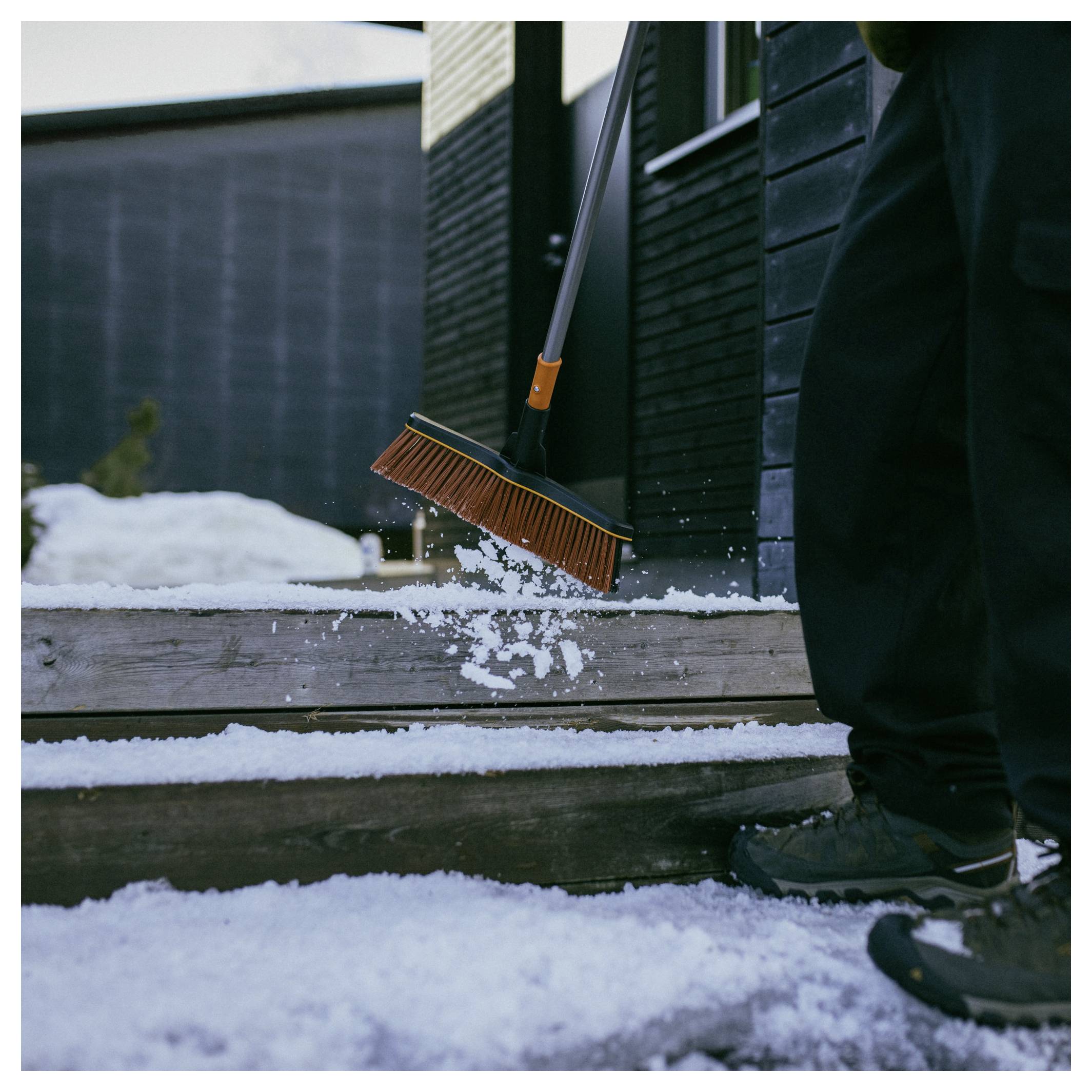 Person in winter clothing clearing snow from steps with an orange broom, outside a dark-colored building.