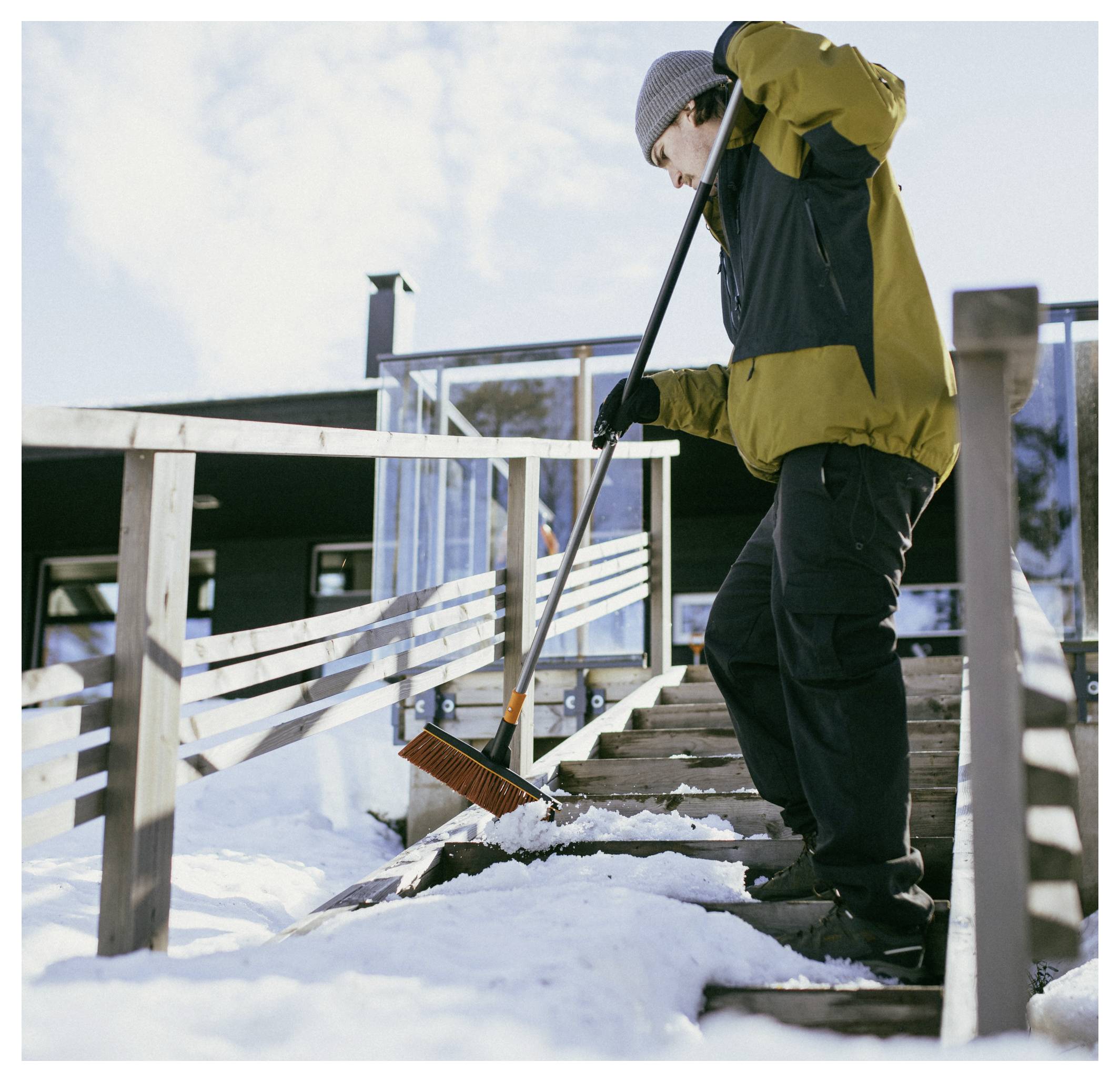 A person in a winter coat and hat shovels snow from wooden stairs leading to a building, with the sun shining overhead.