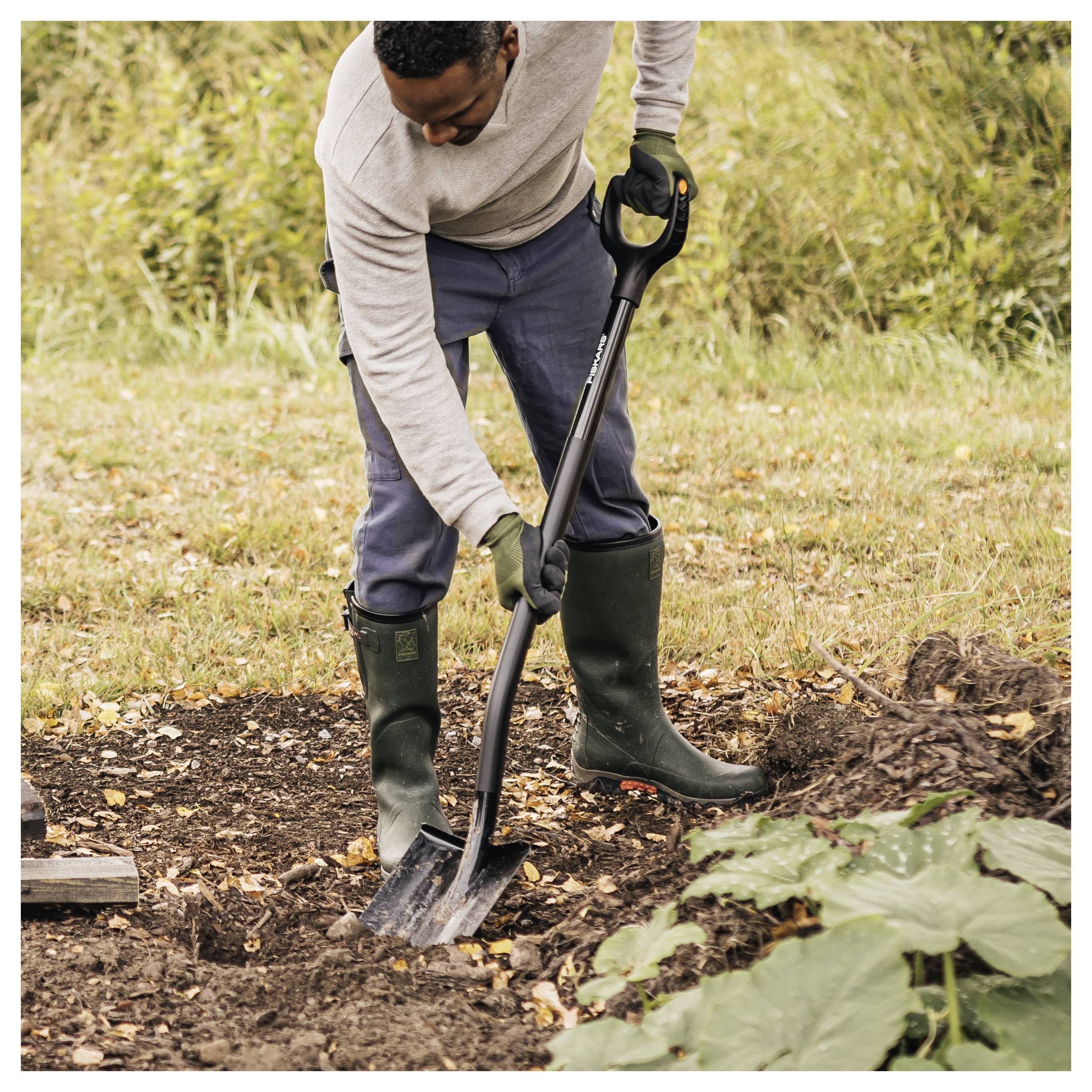 Person digging soil in a garden with a shovel, wearing a grey sweater, blue pants, and green rubber boots, surrounded by greenery.