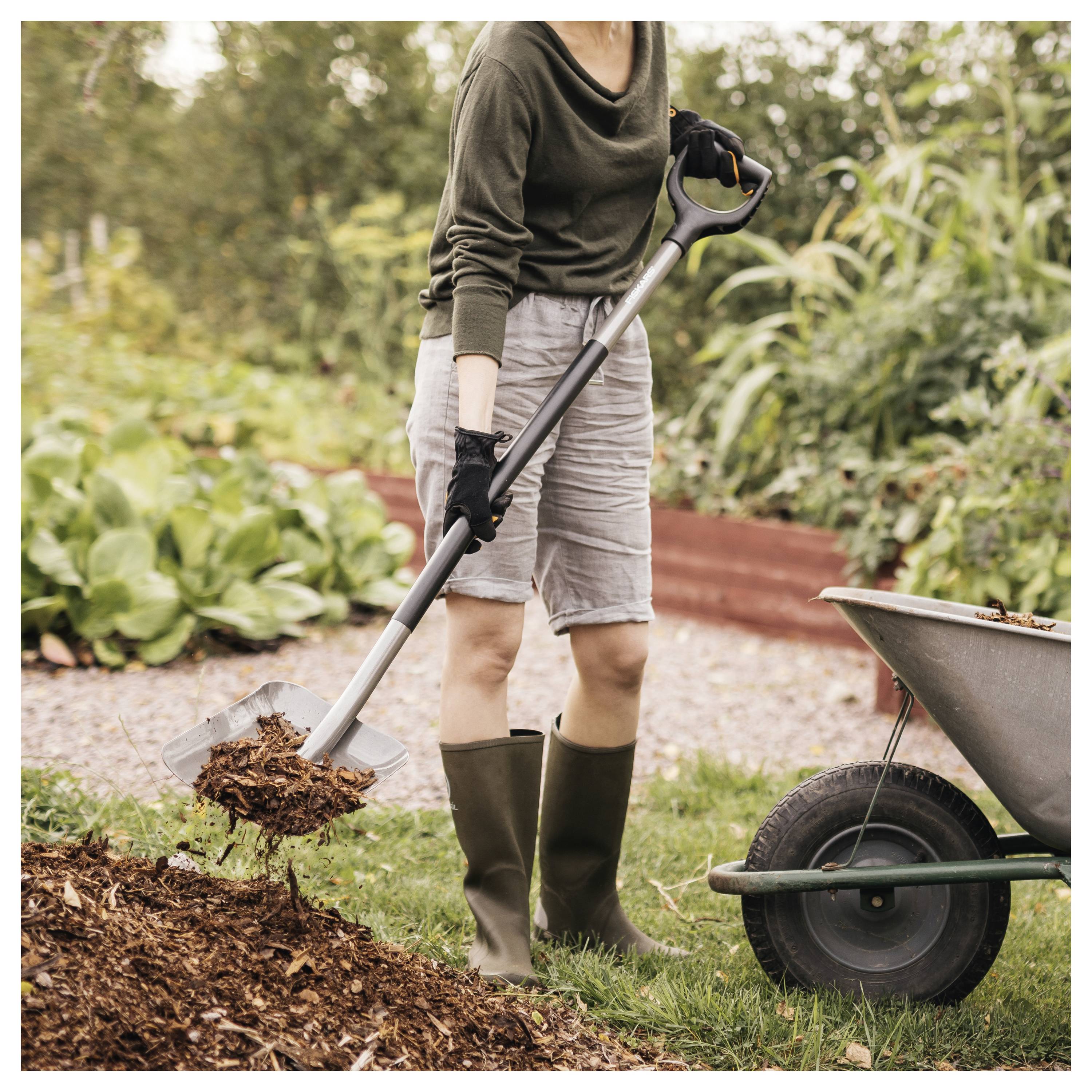 A person in boots and gloves shoveling soil into a wheelbarrow in a garden, surrounded by greenery and plants.