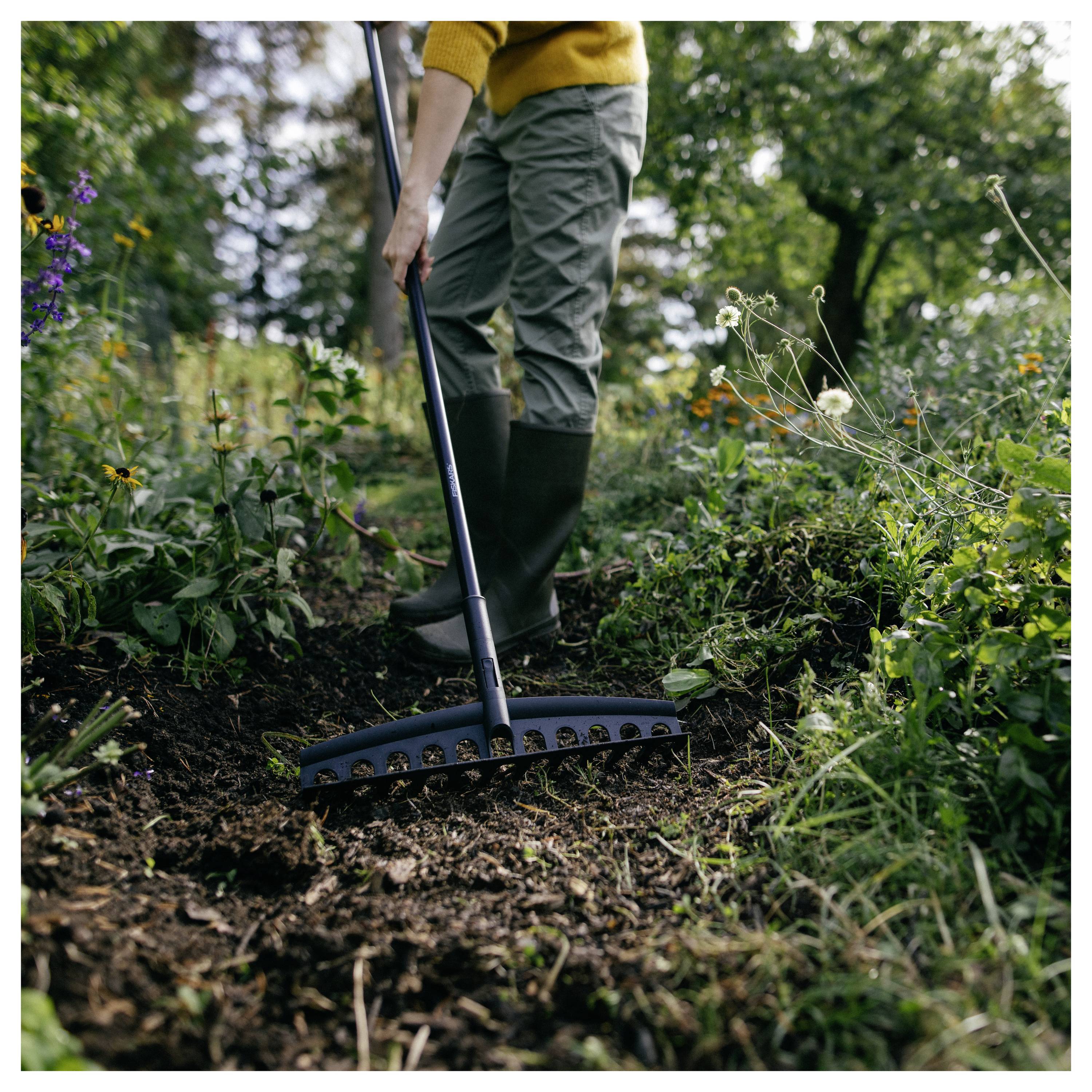 Person in yellow sweater and green pants uses a rake in a garden with flowers and greenery around, suggesting gardening activity.