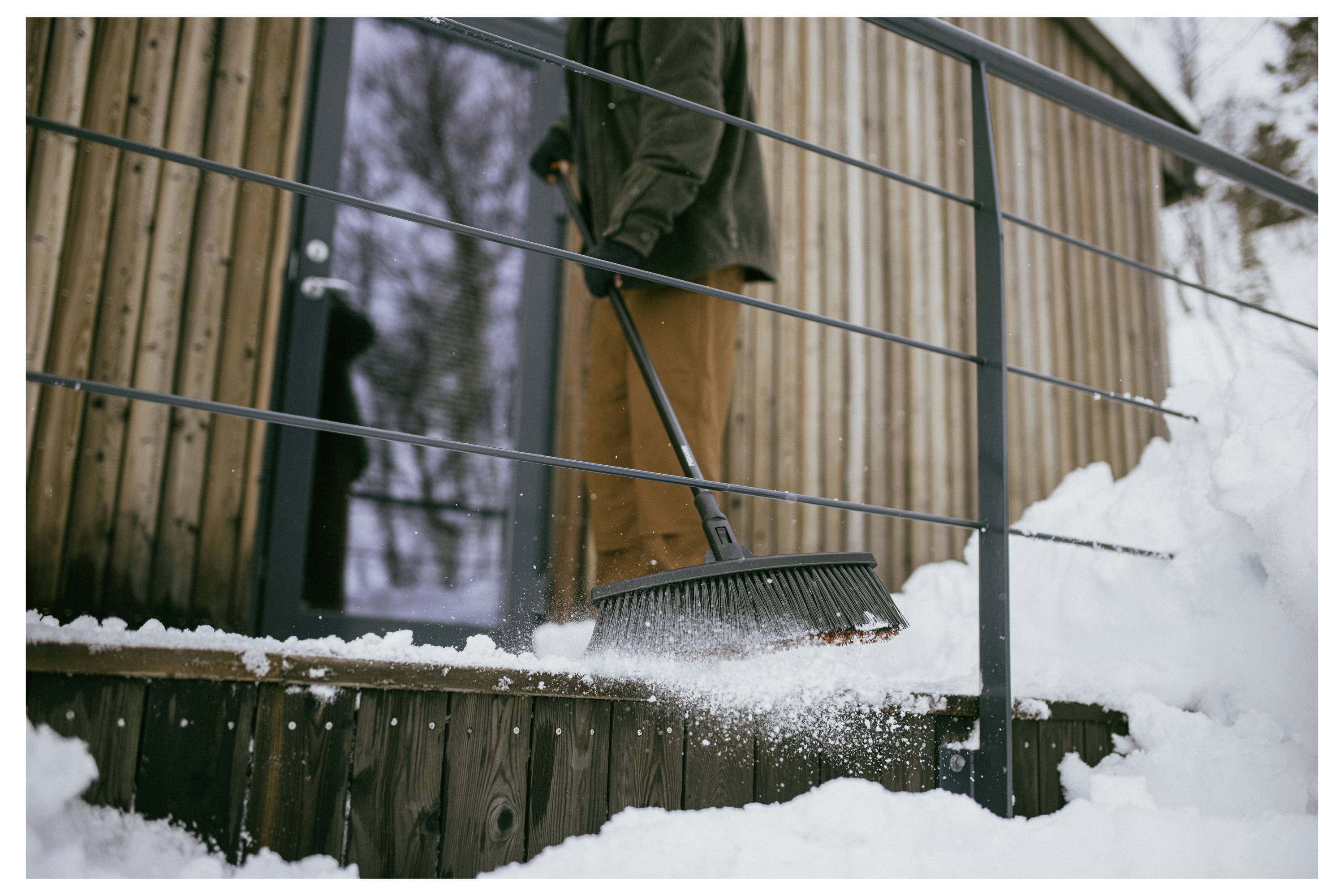 Person sweeping snow off wooden porch steps of a cabin with a broom, wearing a jacket and pants. Snow surrounds the area, trees in background.