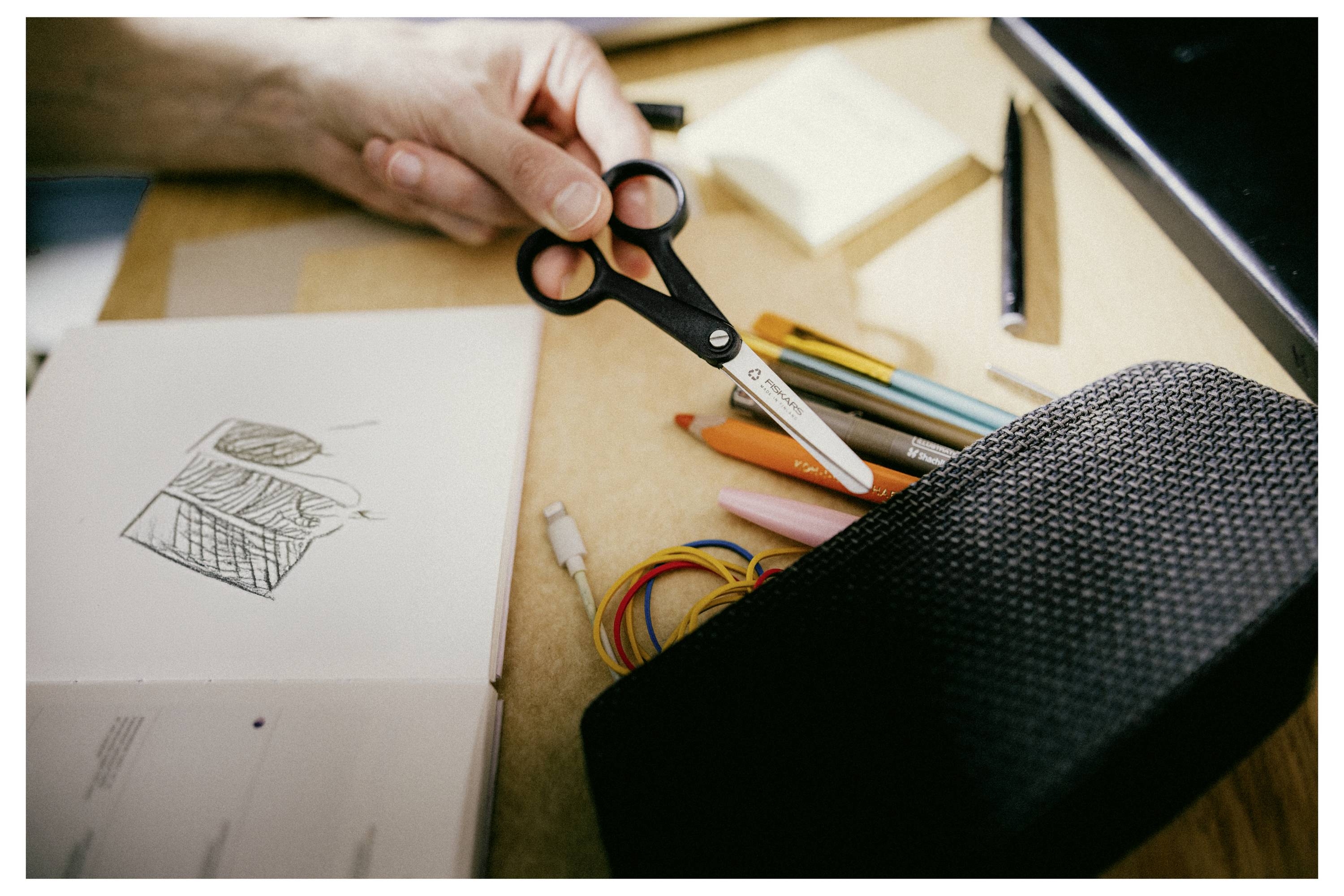 A person's hand holds scissors above a table with a sketchbook, colored pencils, a pencil case, and electrical wires, suggesting art and electronics.