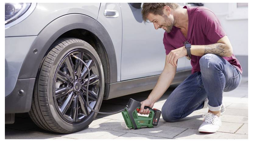 A man kneels next to a car, using a portable air pump to inflate a tire. He wears a maroon shirt and jeans, focused on the task.