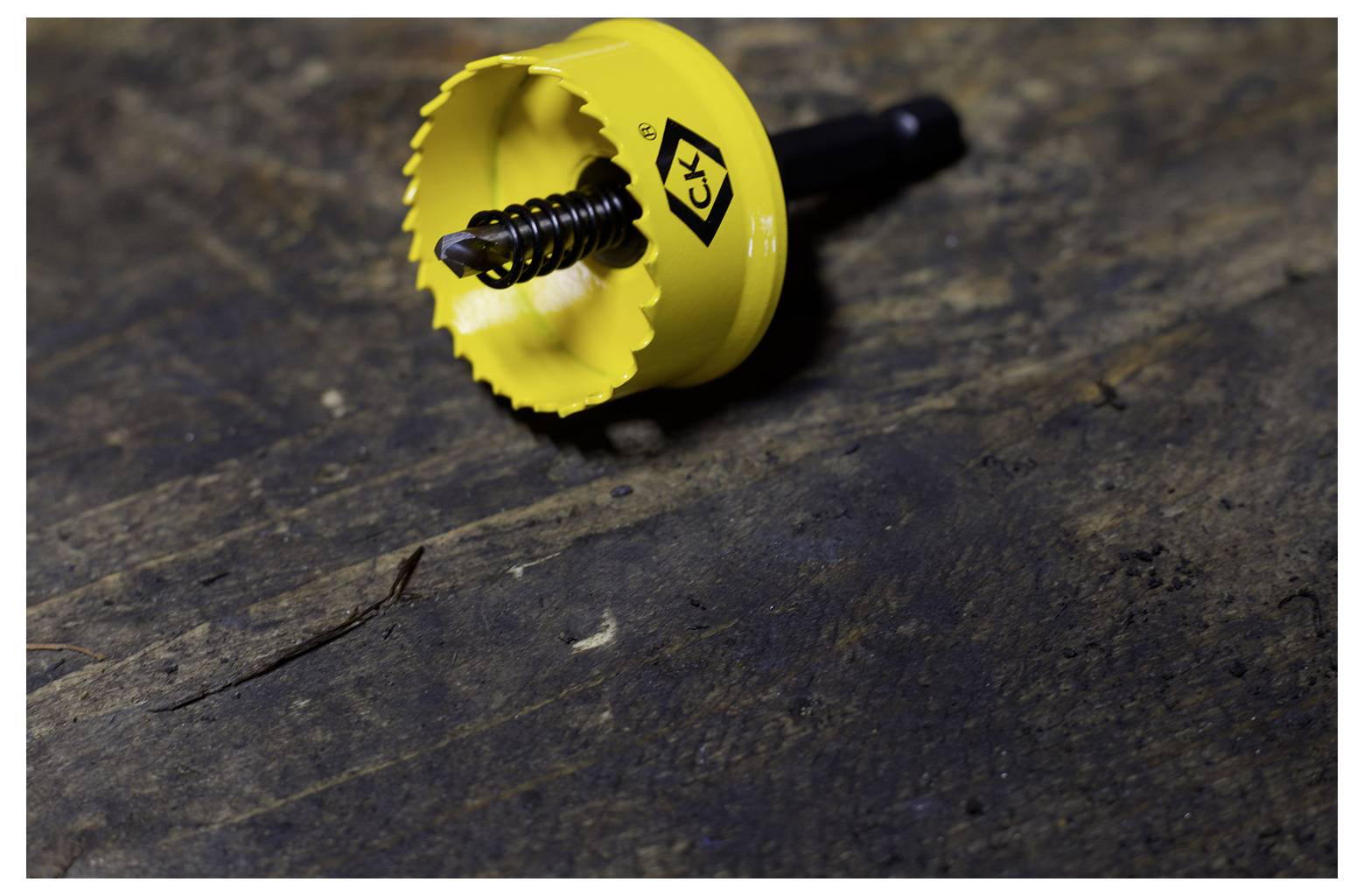 A yellow hole saw drill bit with a metal spring is lying on a dark, textured wooden surface. The tool is designed for cutting circular holes.