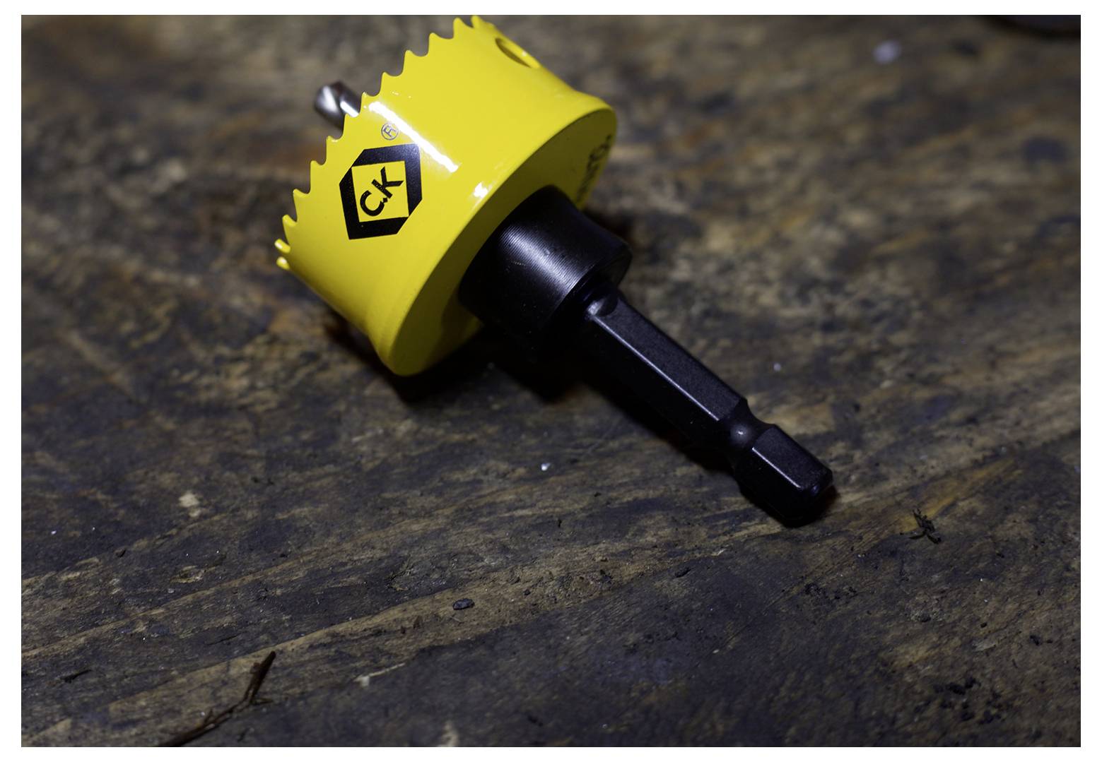 'A C.K. brand yellow hole saw drill bit lies on a dark wooden surface, featuring a jagged edge for cutting circular holes.'
