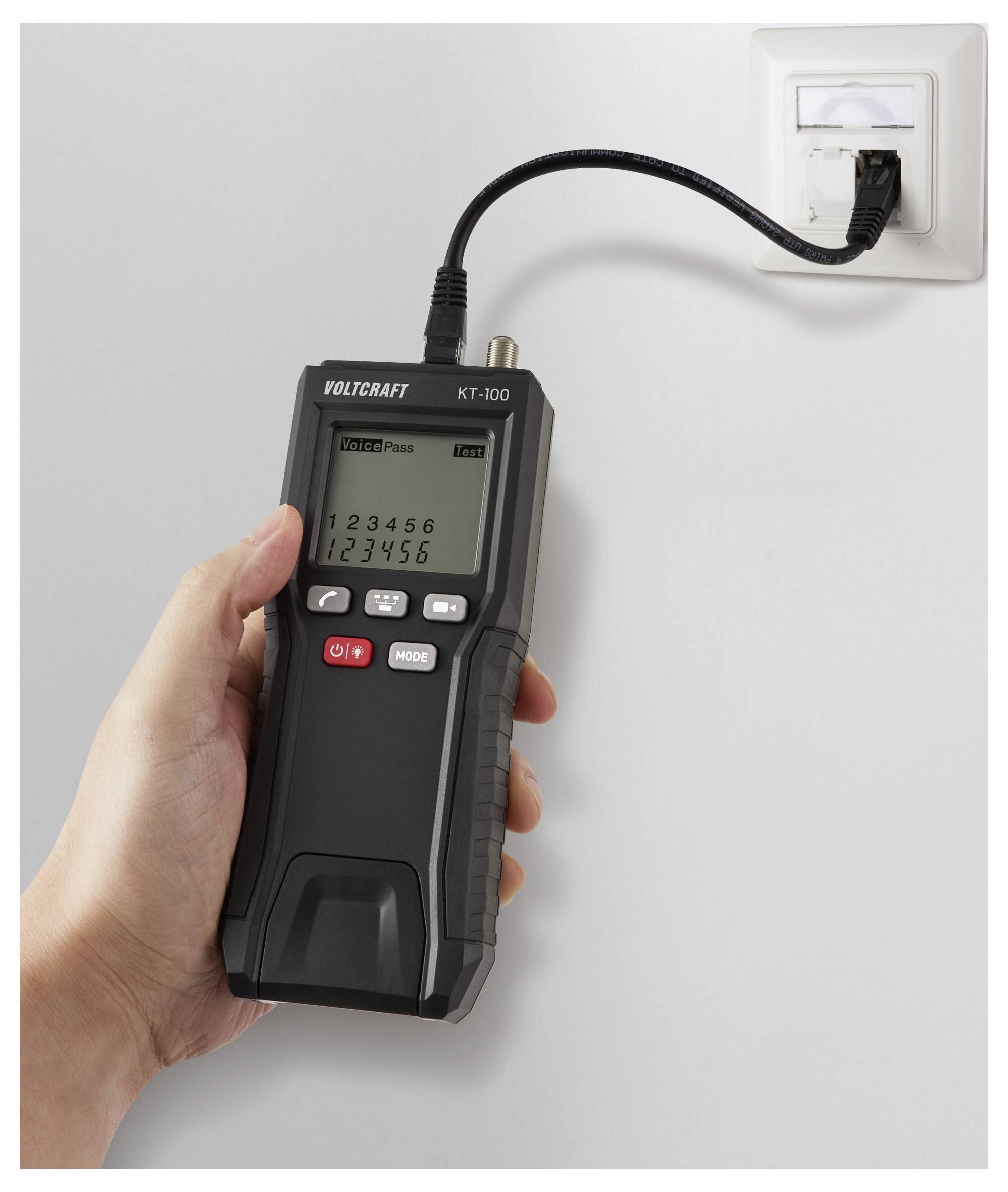 A hand holds a VOLTCRAFT KT-100 cable tester, connected to a wall socket. The display shows numbers and the device is in testing mode.