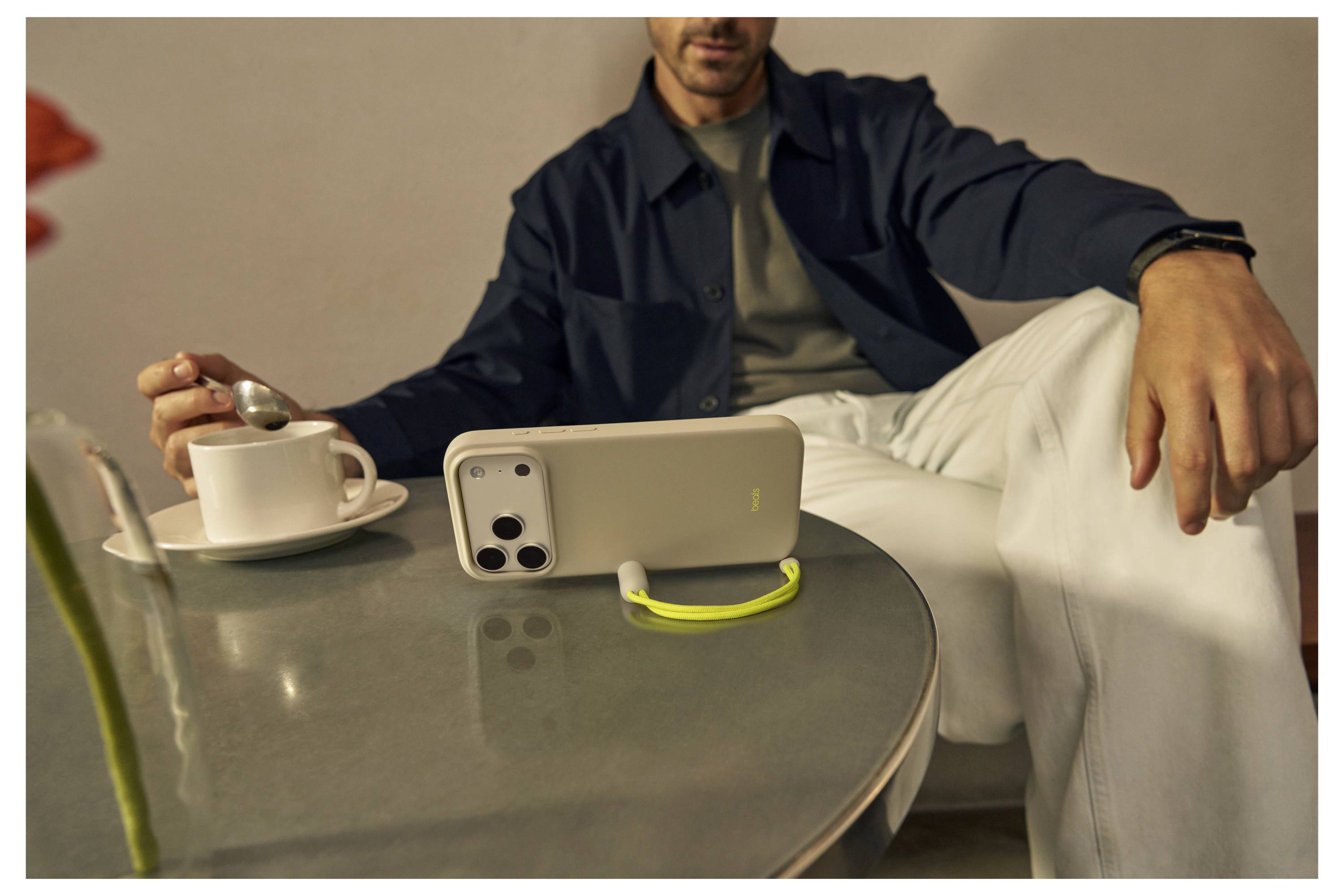 A person sitting at a table with a smartphone propped up, watching or video calling, while holding a spoon and having a cup nearby.
