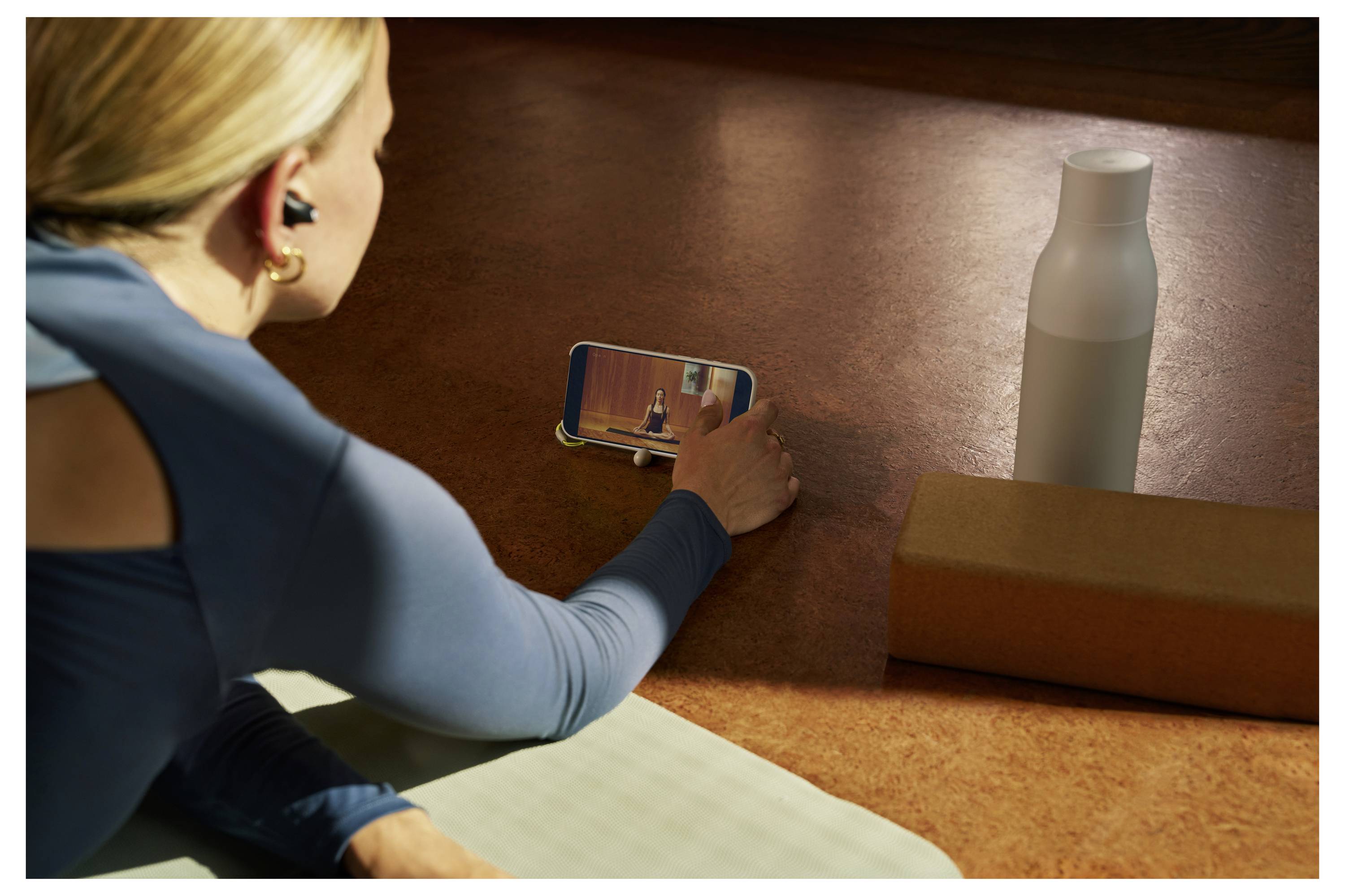 A person wearing headphones watches a yoga video on a phone while lying on a mat, with a bottle and yoga block nearby.