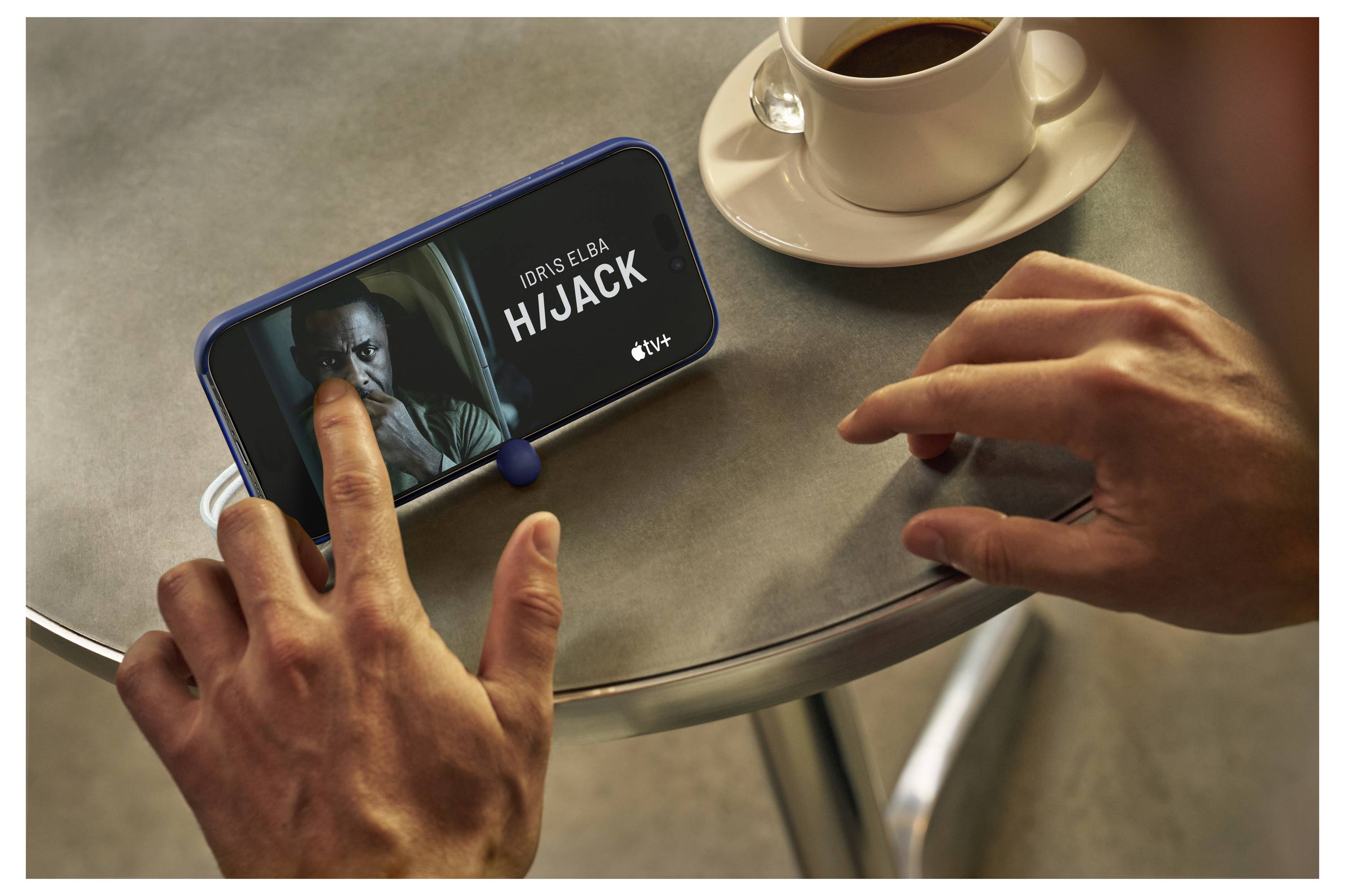 A person interacts with a phone displaying the show 'Hijack' featuring Idris Elba on Apple TV+. A cup of coffee is on the table.