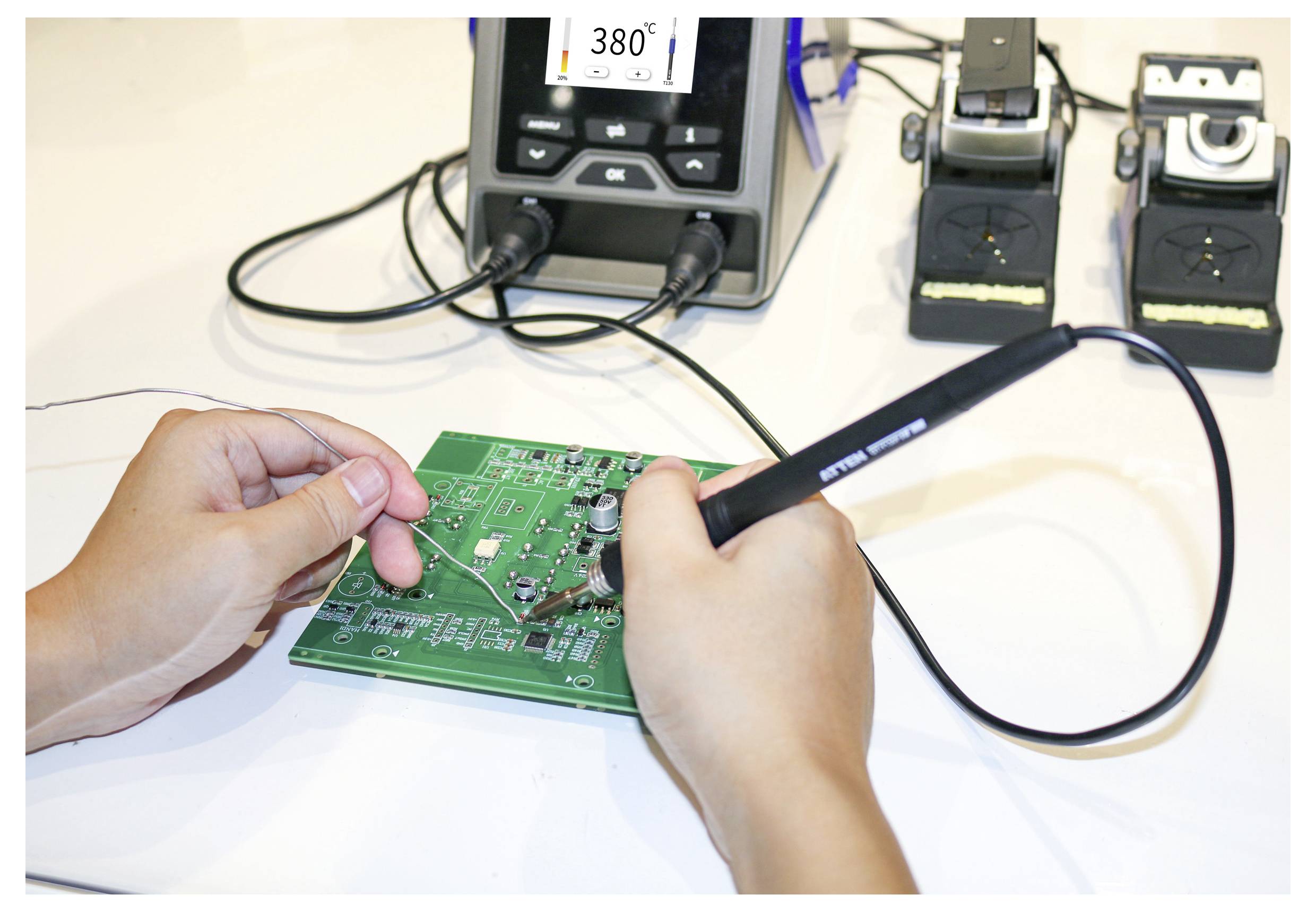 'Hands soldering on a green circuit board with a soldering iron, near a digital soldering station displaying the temperature.'
