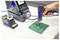 A hand holds a soldering tool applying heat to an electronic circuit board on a work table, with a soldering station and tape dispenser nearby.