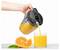 A hand pours orange juice from a pitcher into a glass on a white surface. Fresh orange slices and leaves are next to the glass.