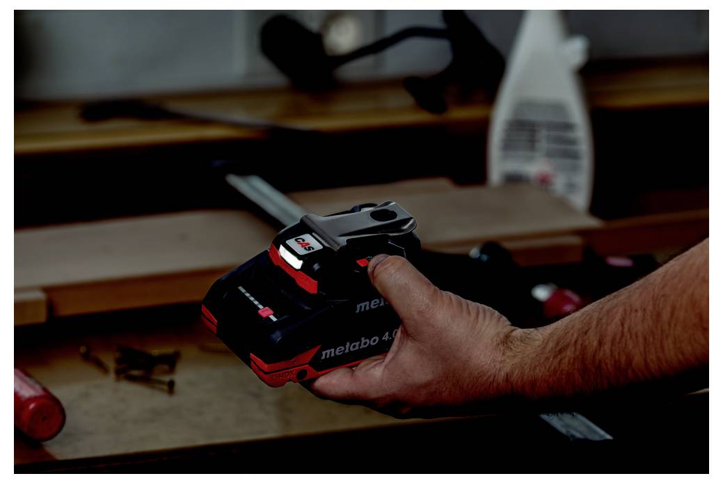 A person holds a 'metabo 4.0' battery pack in a workshop setting, with tools and wooden boards in the background.