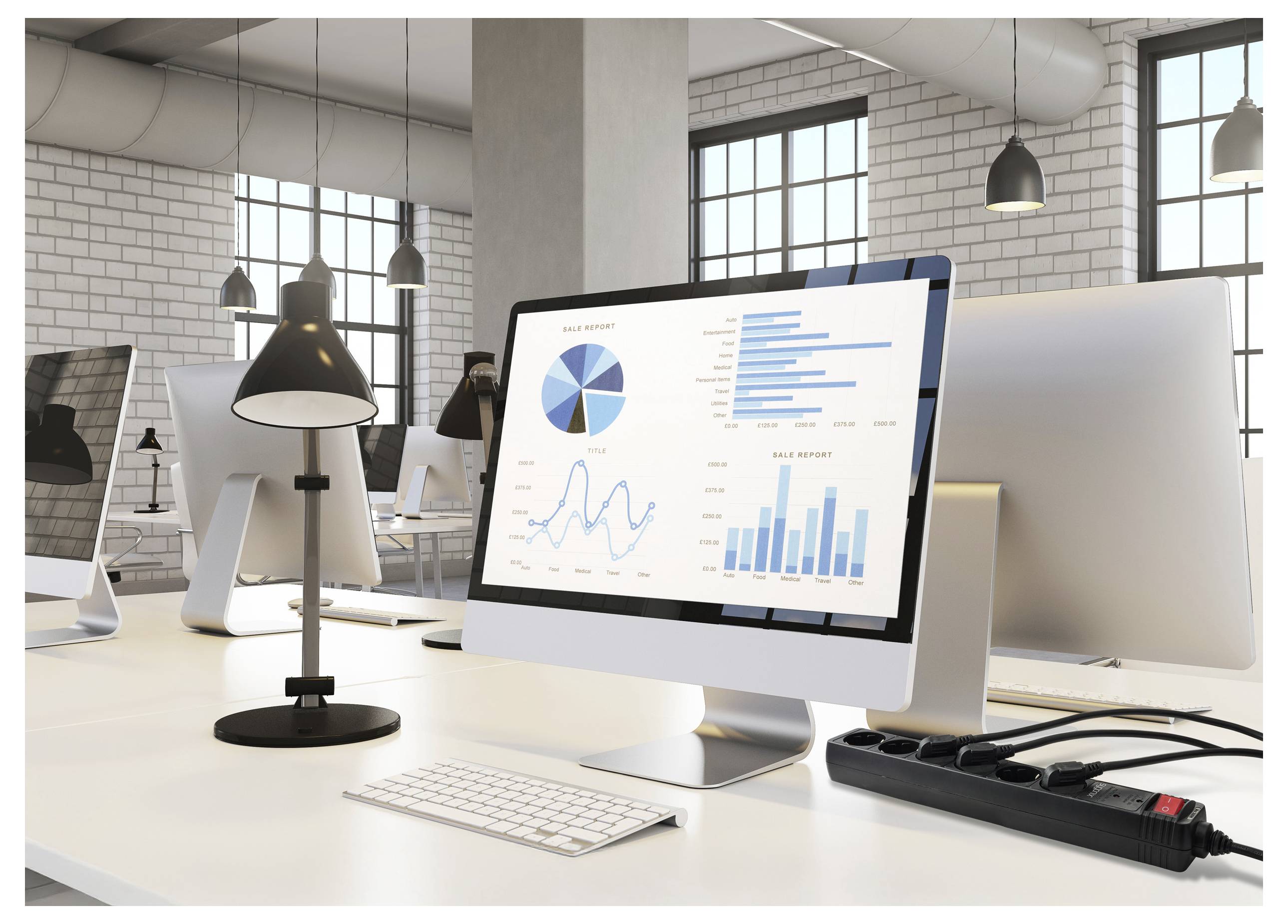 A modern office workspace with a computer displaying sales reports and graphs. The surroundings include white brick walls and industrial-style windows.