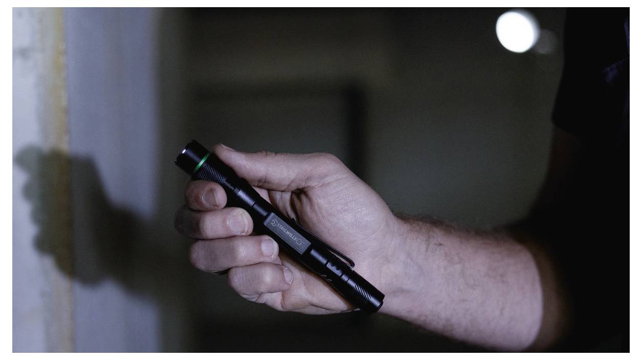 A person holds a small flashlight, illuminating a wall in a dimly lit environment.