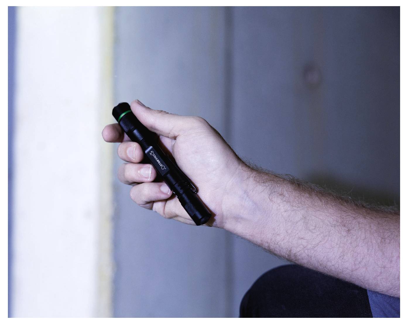 A person holding a black flashlight with a green button, illuminating a dimly lit area.
