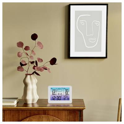 A digital clock on a wooden table shows '12:30' with additional information such as date, temperature, and humidity. Nearby is a vase with branches and a framed abstract face art on the wall.