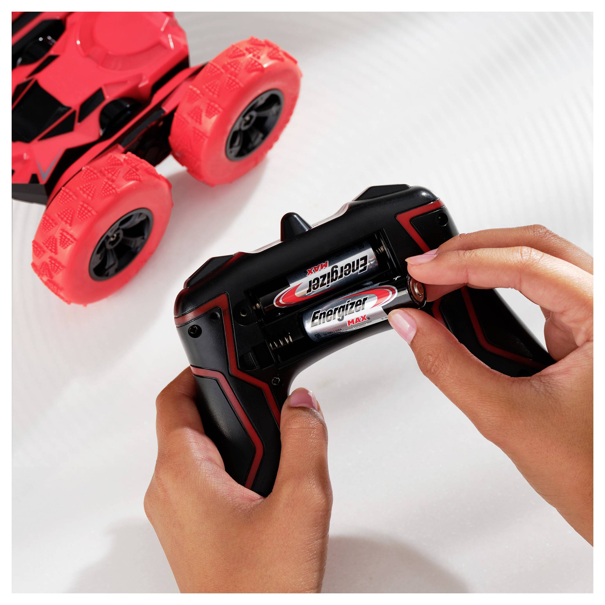 A person is placing batteries into a remote control for a toy car. The toy car has large, red wheels.