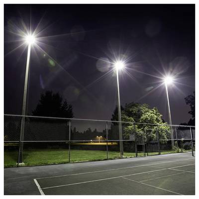 A tennis court at night is illuminated by three tall floodlights, casting bright light over the fenced area. Trees are visible in the background.