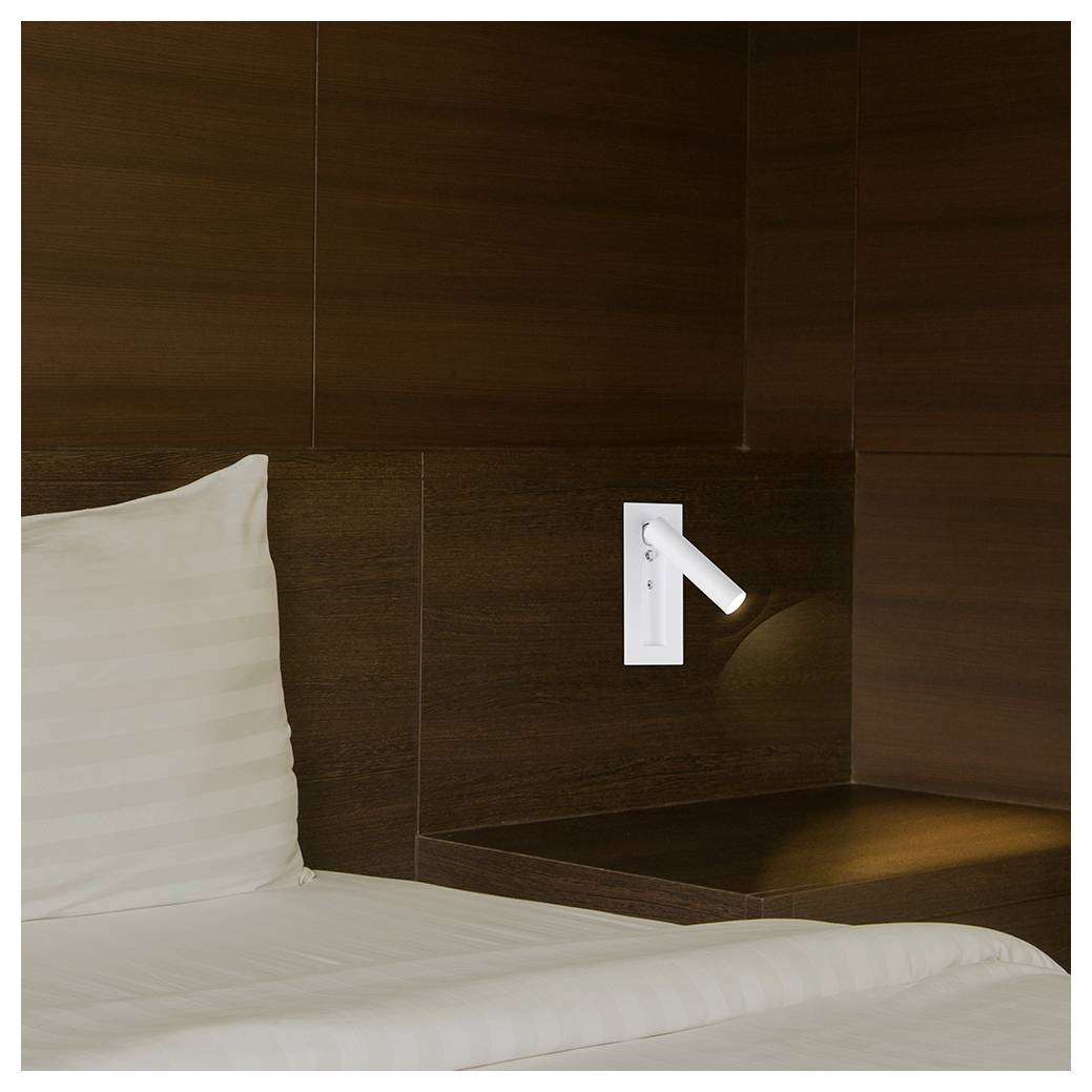 A modern hotel room corner with dark wood paneling, a mounted white reading light, and part of a white pillow on the bed.