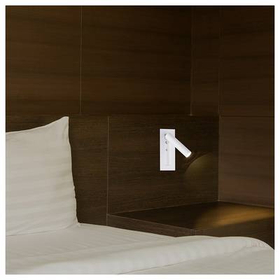 A modern hotel room corner with dark wood paneling, a mounted white reading light, and part of a white pillow on the bed.