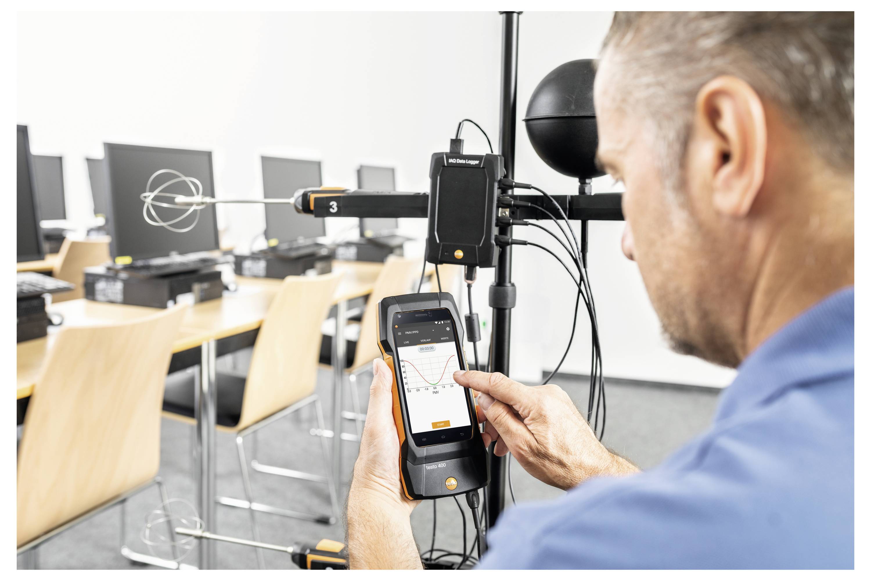 A man in a blue shirt uses a handheld device to measure sound or signal levels in a computer lab with multiple monitors.