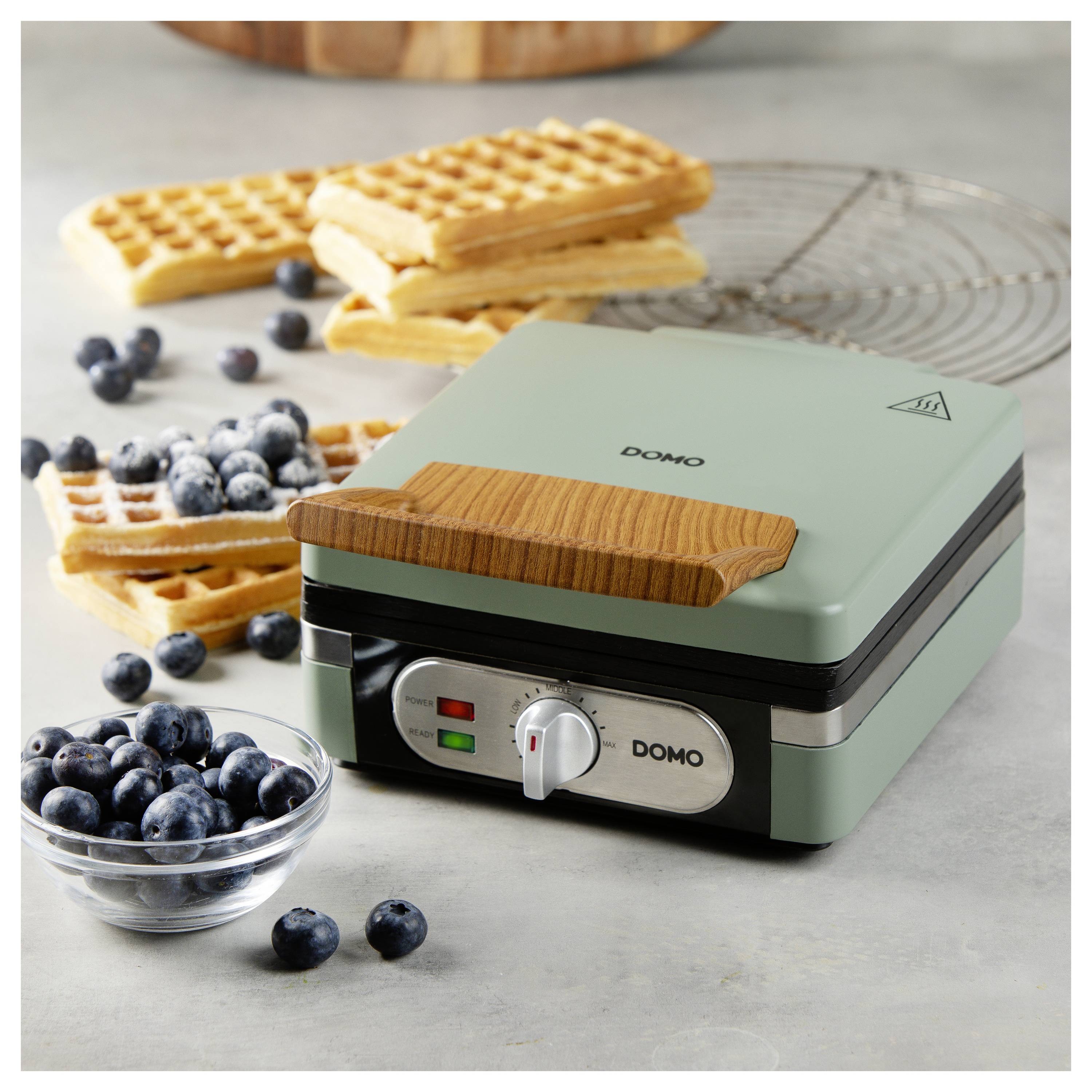 A waffle maker with a wooden handle and control dial, surrounded by freshly made waffles and a bowl of blueberries on a countertop.
