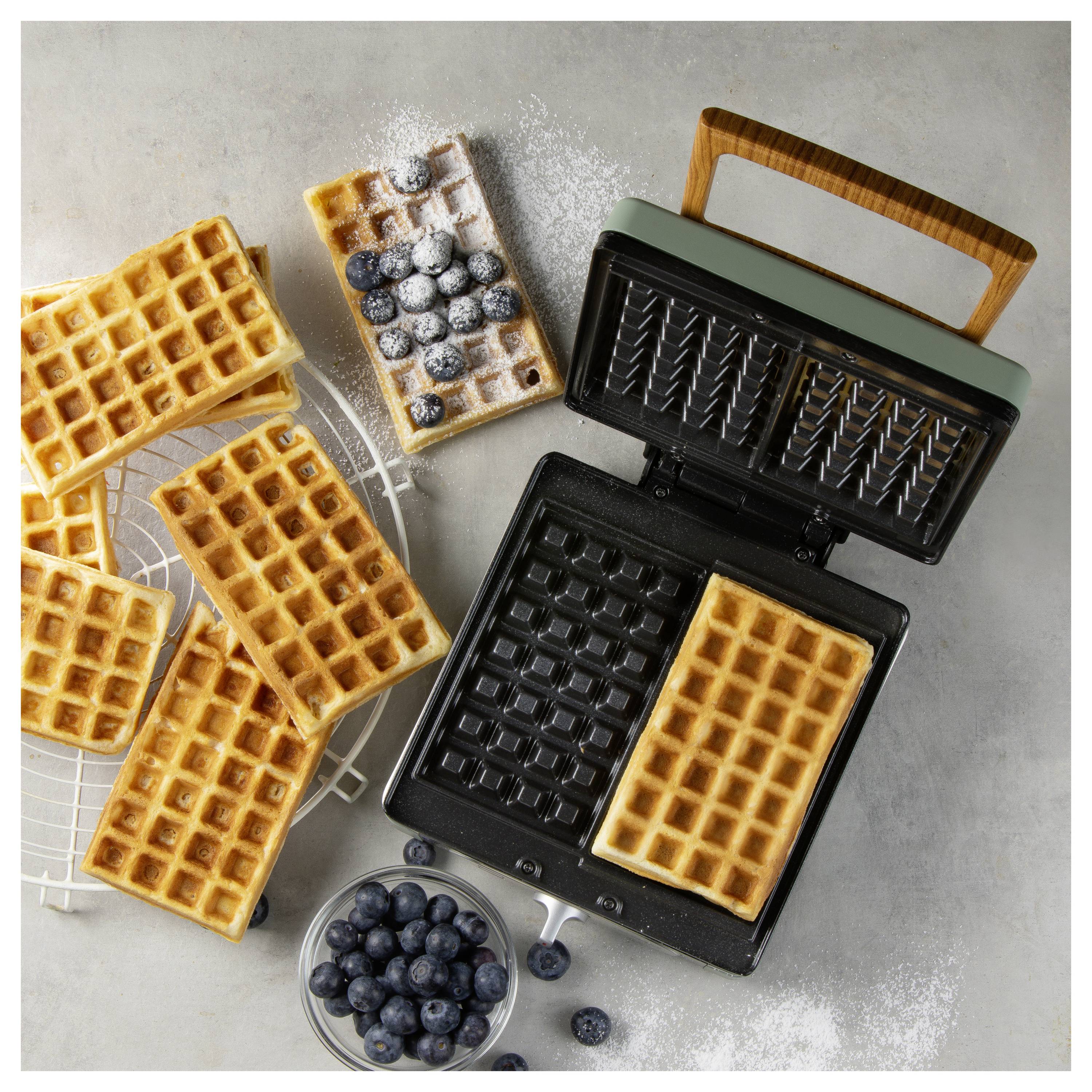 Waffles and blueberries with a waffle maker. Waffles lay on a cooling rack and one inside the open waffle maker; a bowl of blueberries nearby.