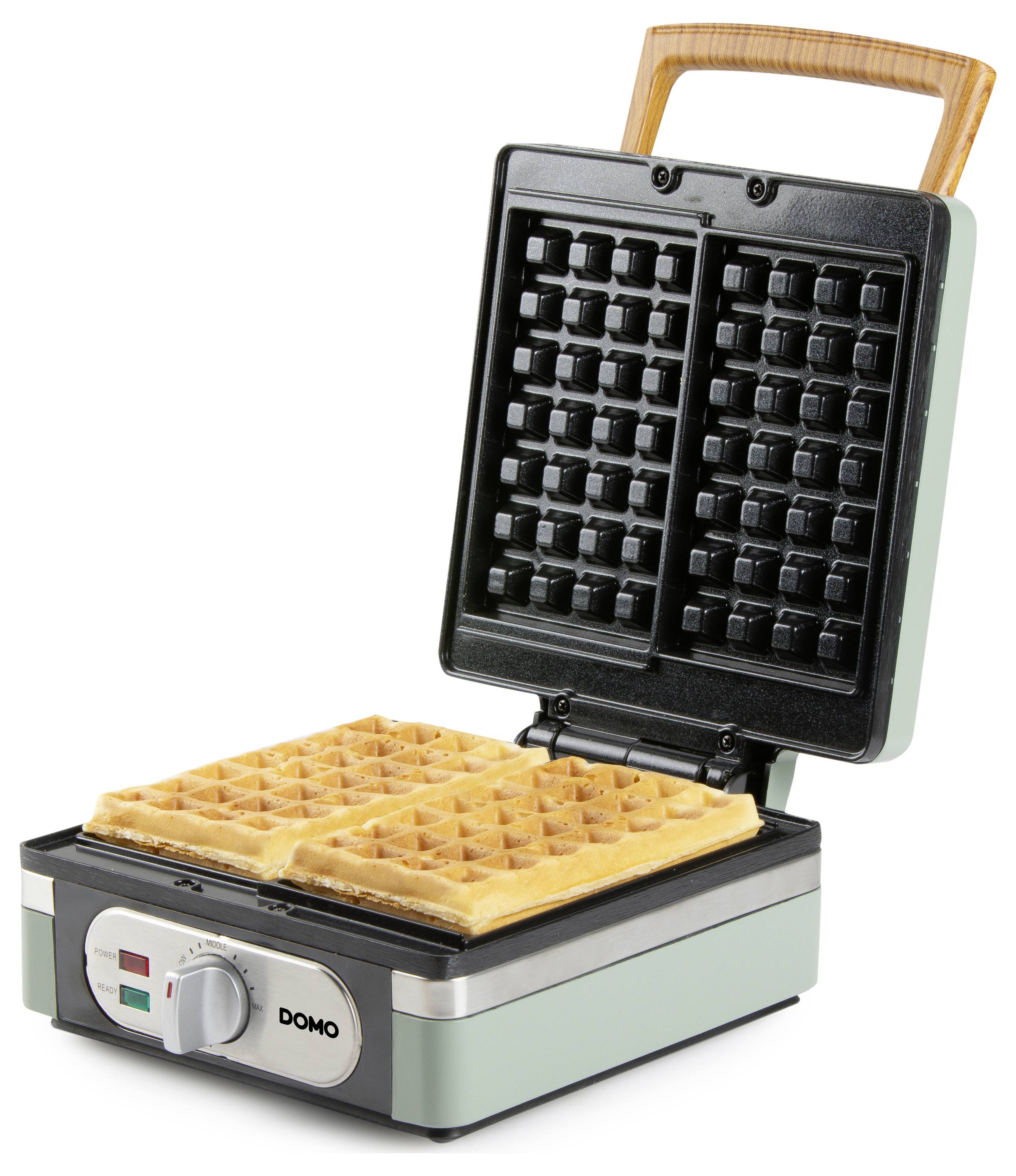 A waffle maker with an open lid showing two square waffles inside, ready to be served. The device is green with a wooden handle.