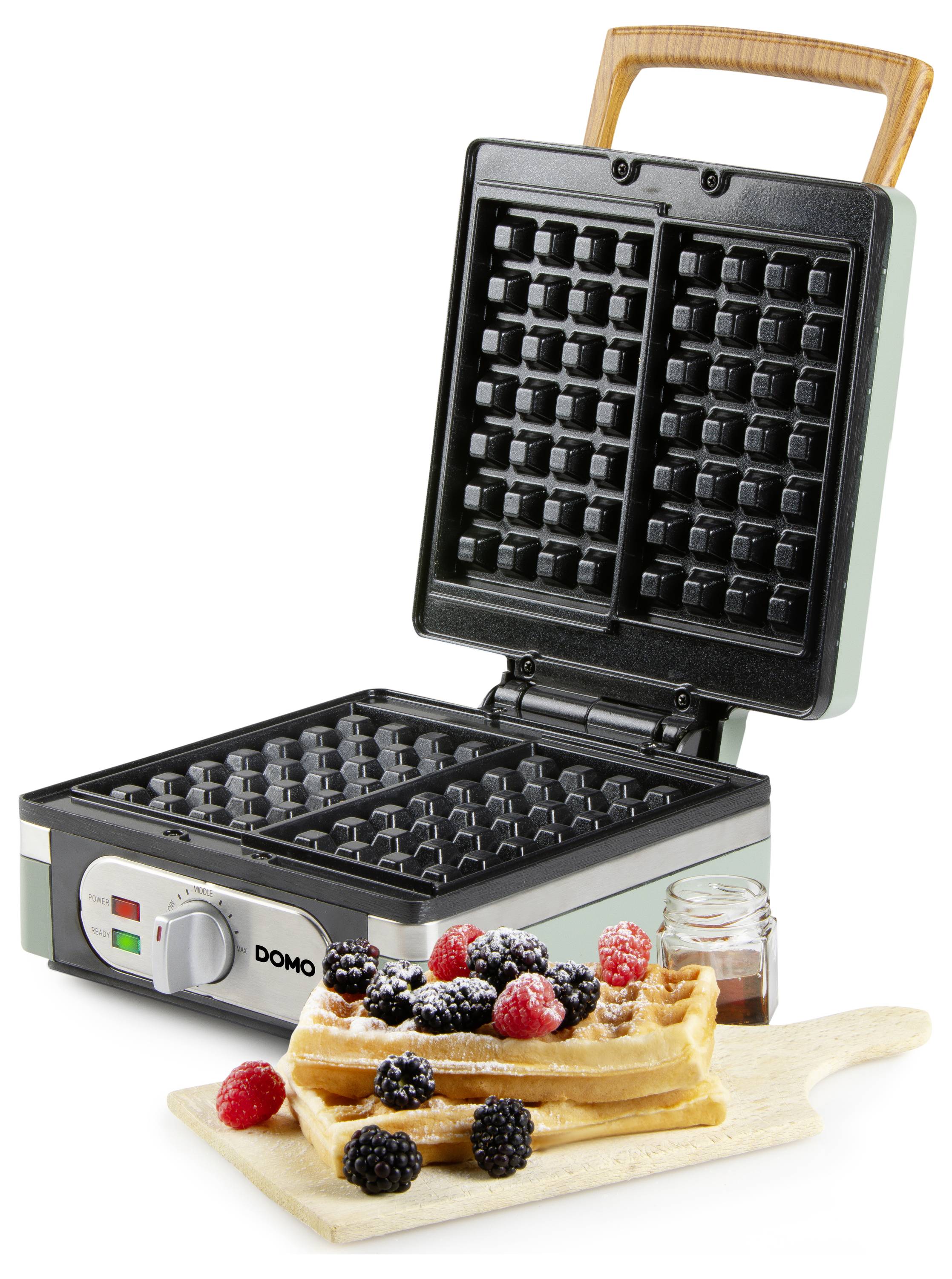 A waffle iron is open with freshly baked waffles on a wooden board topped with berries, next to a small jar of syrup.