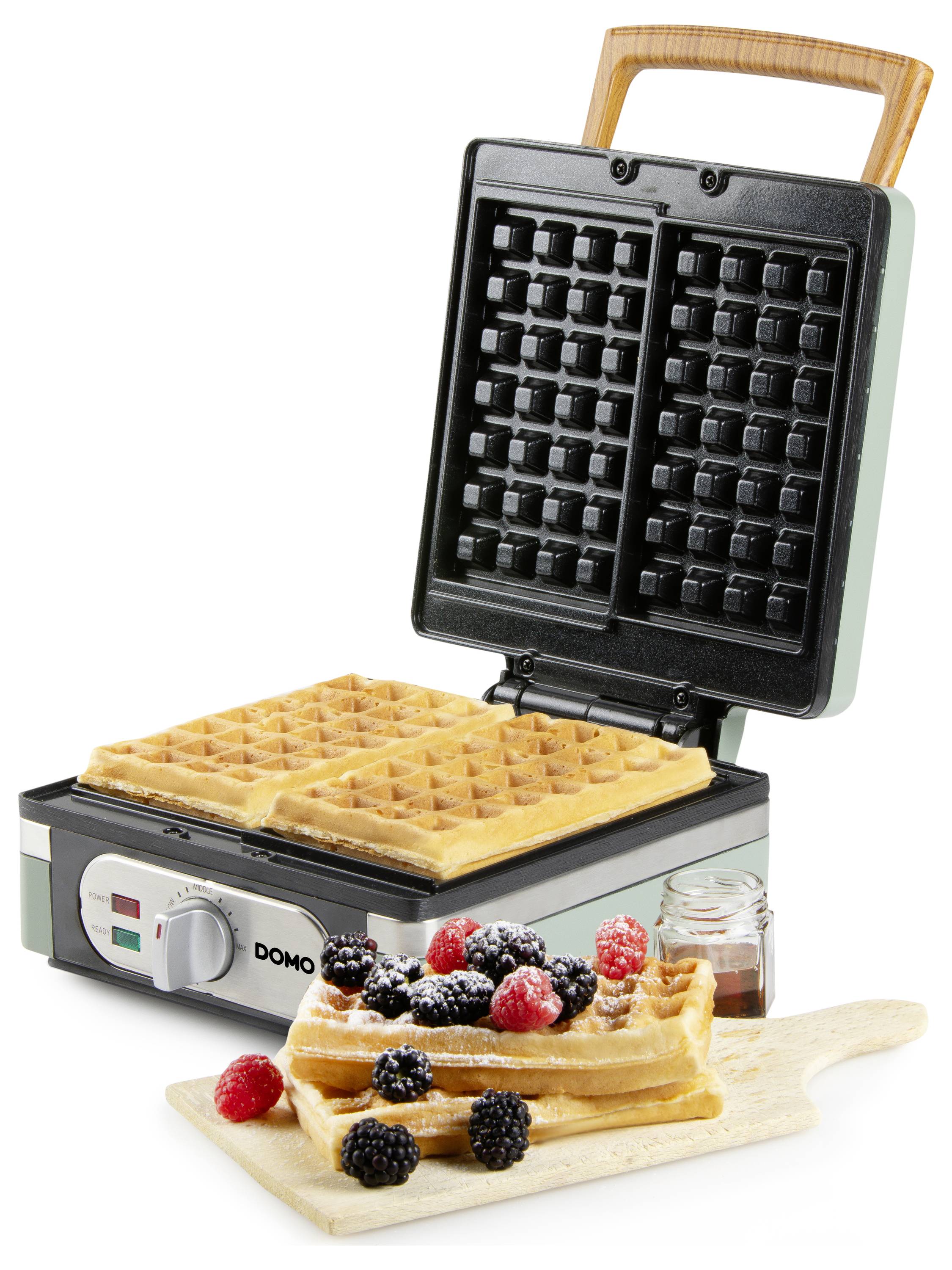 A waffle maker with two waffles inside, next to a wooden board with stacked waffles garnished with strawberries, raspberries, and blackberries.