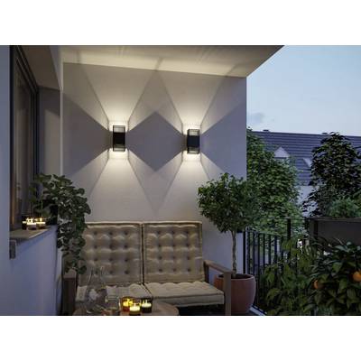 A cozy balcony scene at dusk with two lit wall sconces casting light patterns, a cushioned sofa, small table, and potted plants.