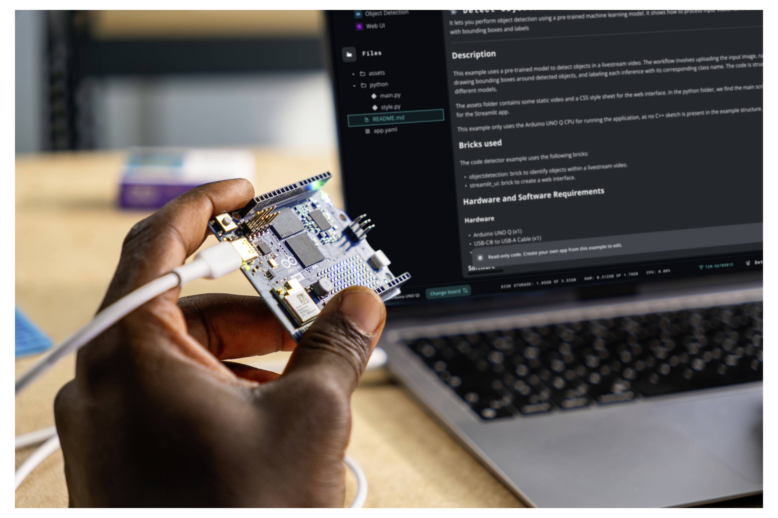 A person holds a microcontroller board connected by a cable in front of a laptop displaying coding instructions.