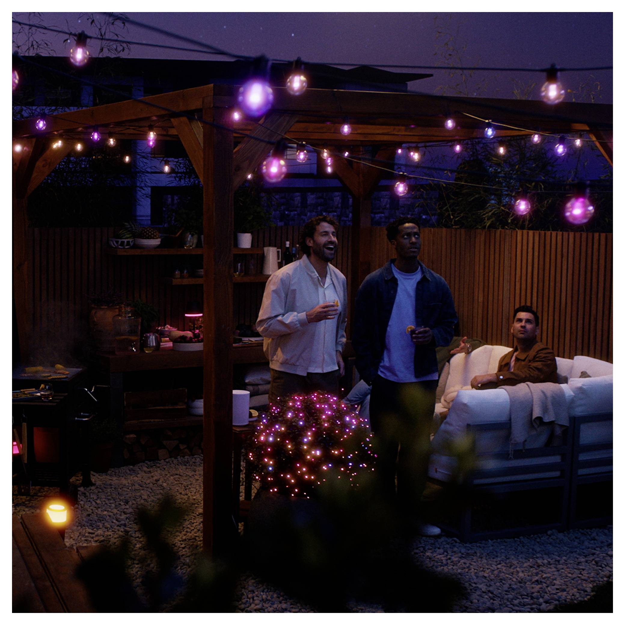 Three people enjoy a relaxed evening under a pergola with purple string lights. One is sitting on a couch, holding a drink.
